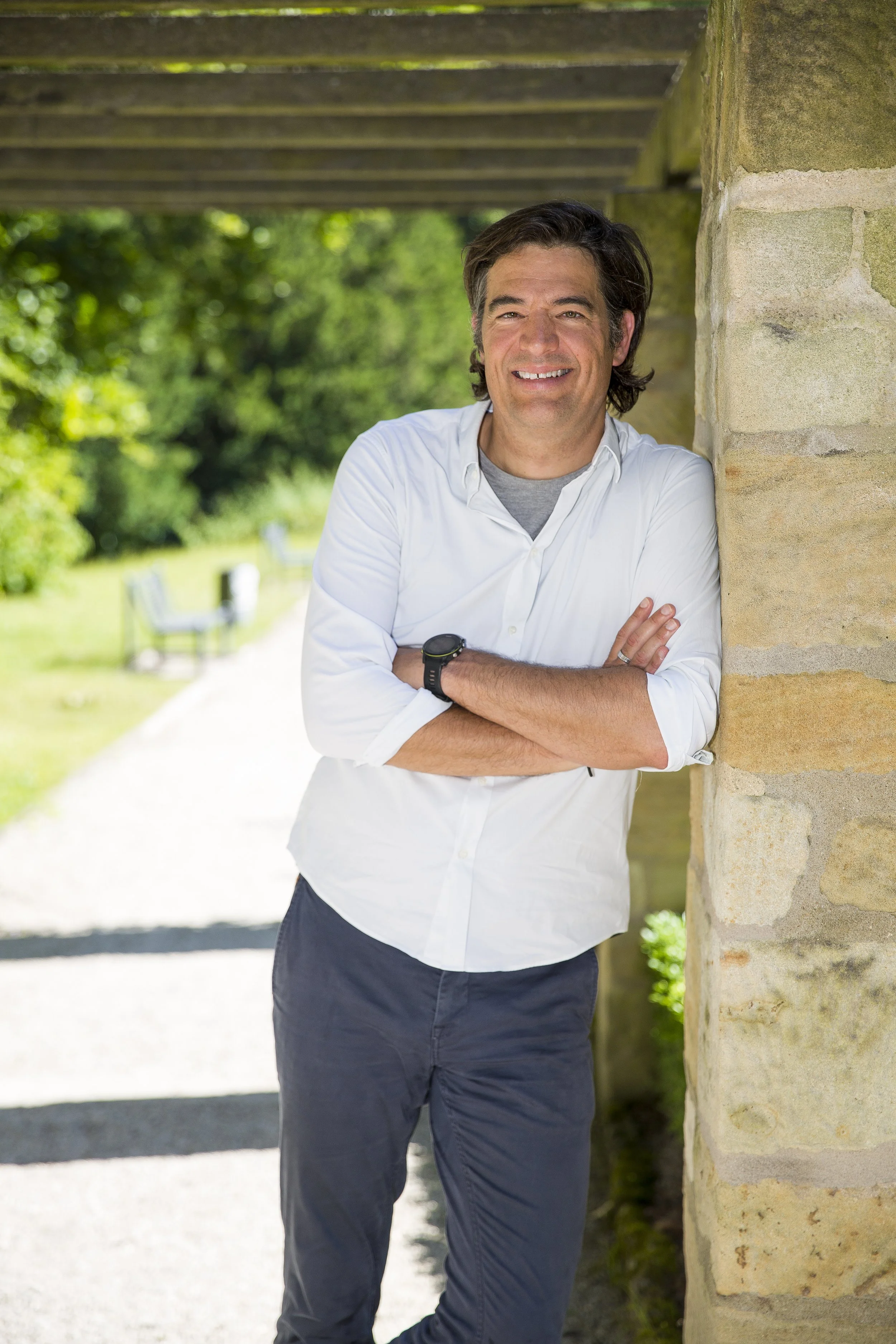 Ein lächelnder Mann in weißem Hemd und dunklen Hosen lehnt an einer Steinmauer im Freien, umgeben von grünen Bäumen und Bänken.