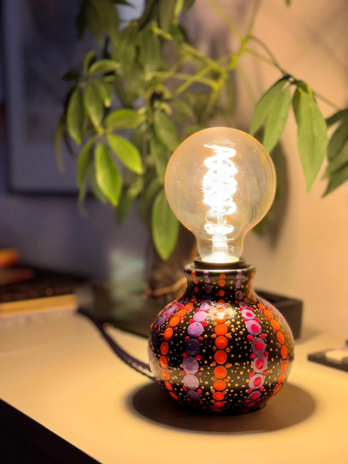 Decorative table lamp with a painted black base featuring multicolored dots and a clear incandescent bulb, with green leaves of a plant in the background.