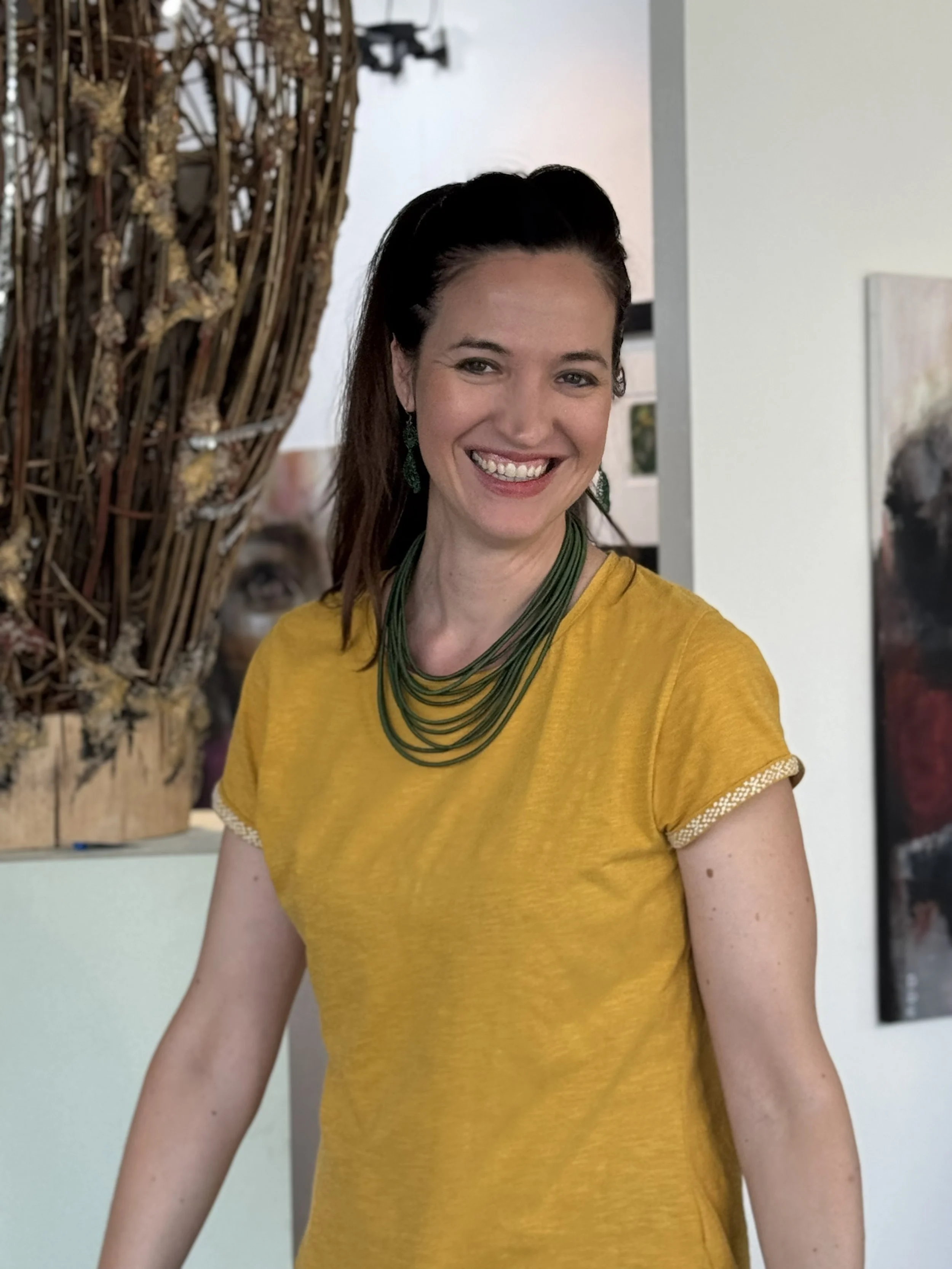 A smiling woman with dark hair and wearing a yellow top and green jewelry, standing indoors with artwork and a sculpture in the background.