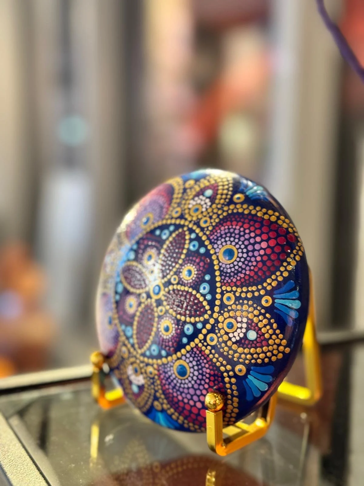 Decorative painted stone with dot patterns in yellow, pink, blue, and purple, displayed on a small gold stand.