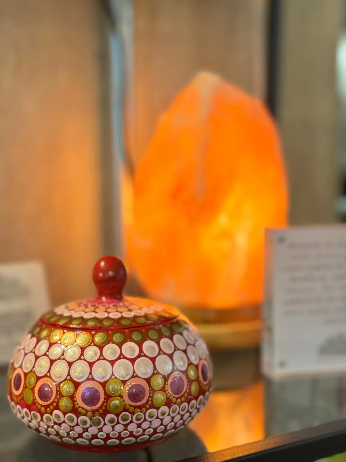 A decorative glass orb with red, white, green, and gold polka dot pattern in front of a lit salt lamp in a display case.