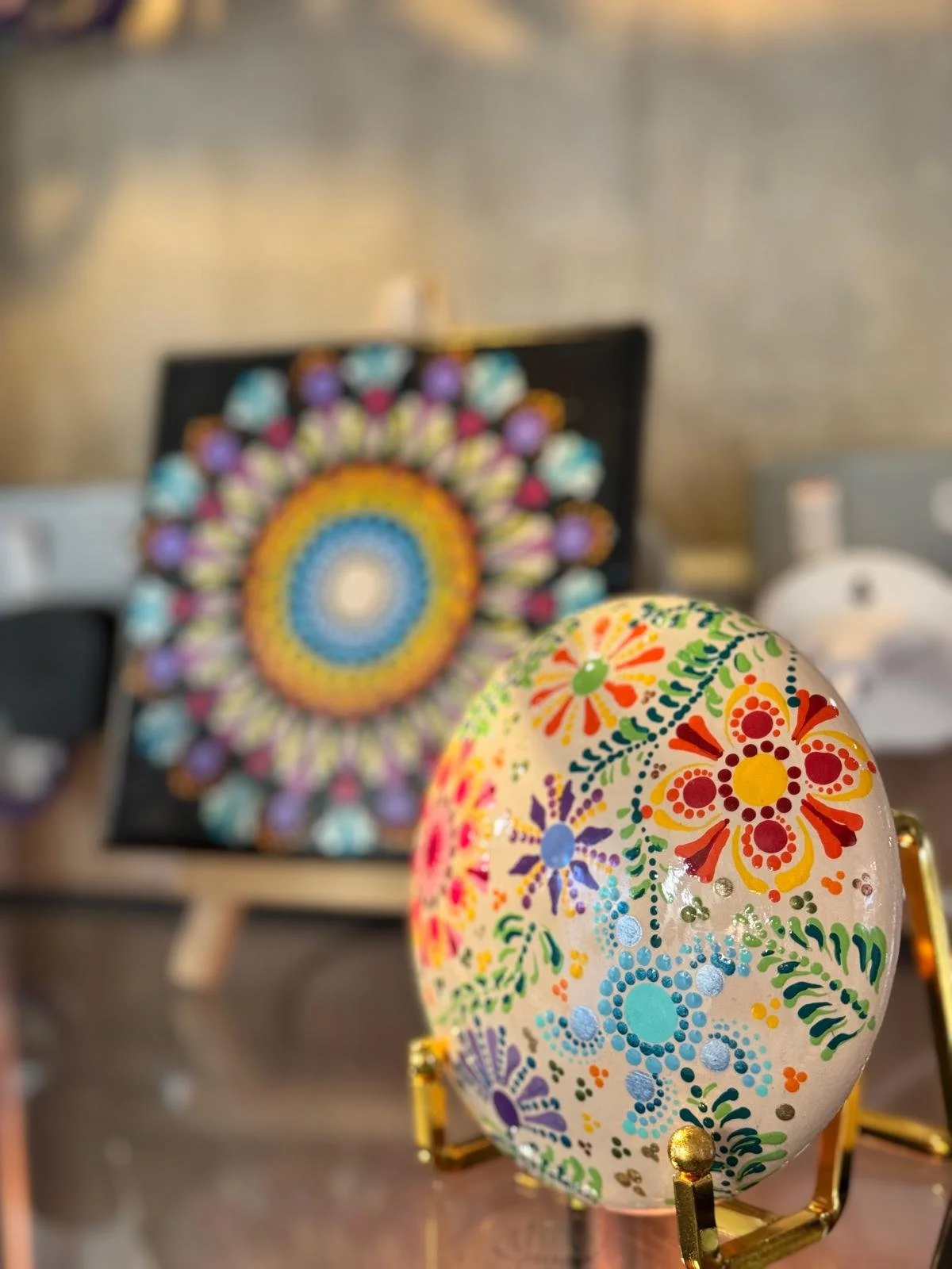 Decorative painted egg on gold stand in foreground, blurred colorful mandala art in background.