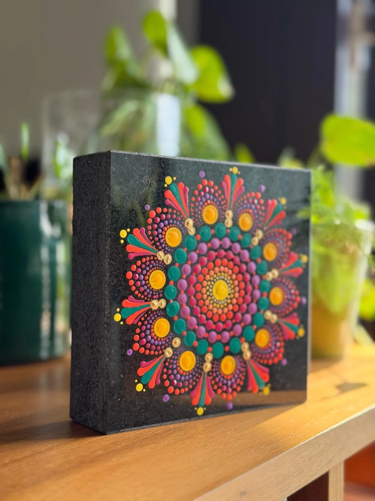 A black cube with colorful dot and line mandala art on its front face, sitting on a wooden surface, with potted plants in the background.