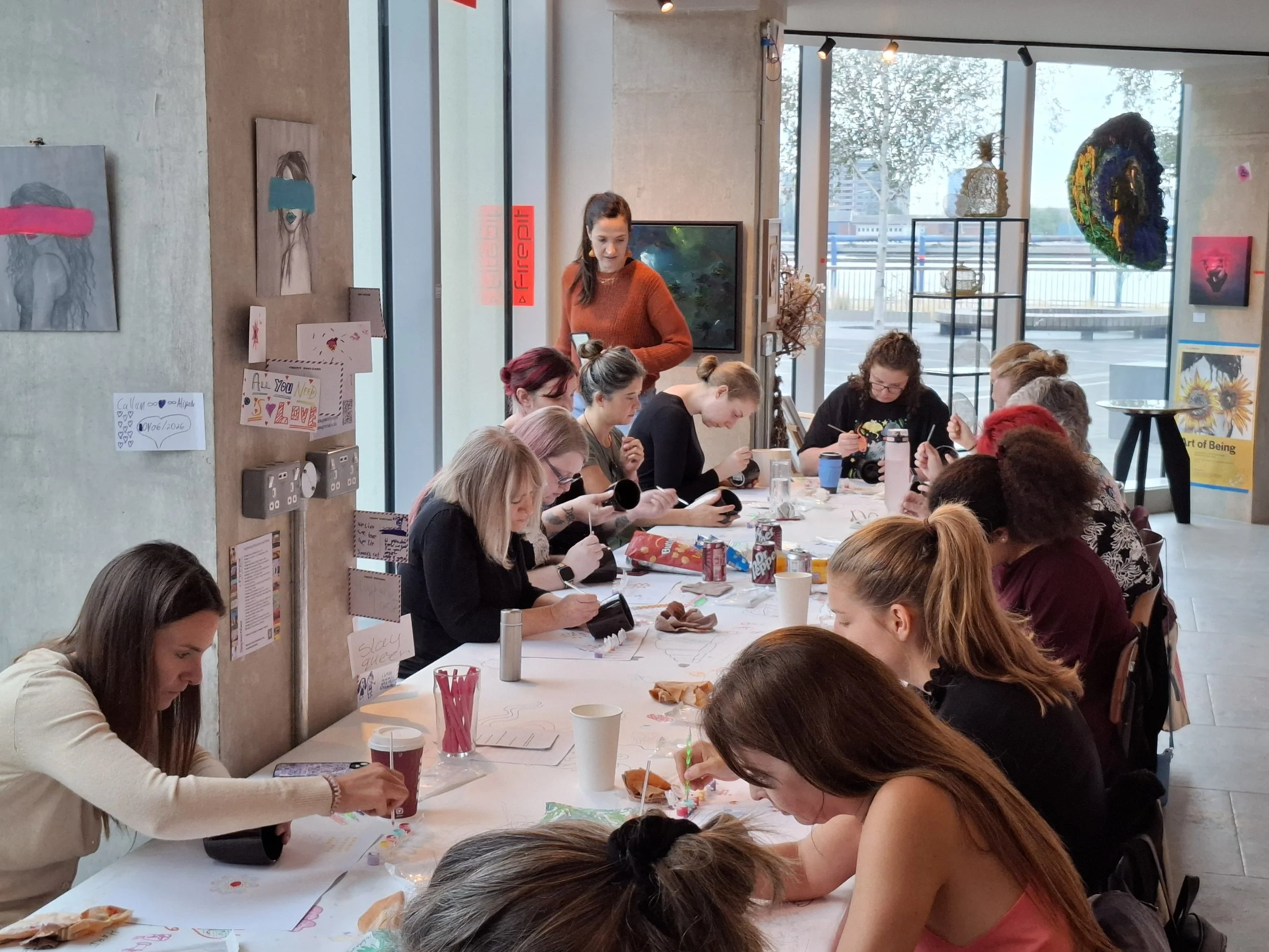 People sitting around a large table engaging in arts and crafts in a bright, modern art gallery with artwork and large windows in the background.