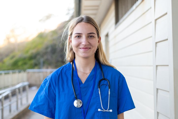 Young female nurse outdoors wearing blue scrubs and a stethoscope, smiling in front of a light-colored building with a fence and greenery in the background.