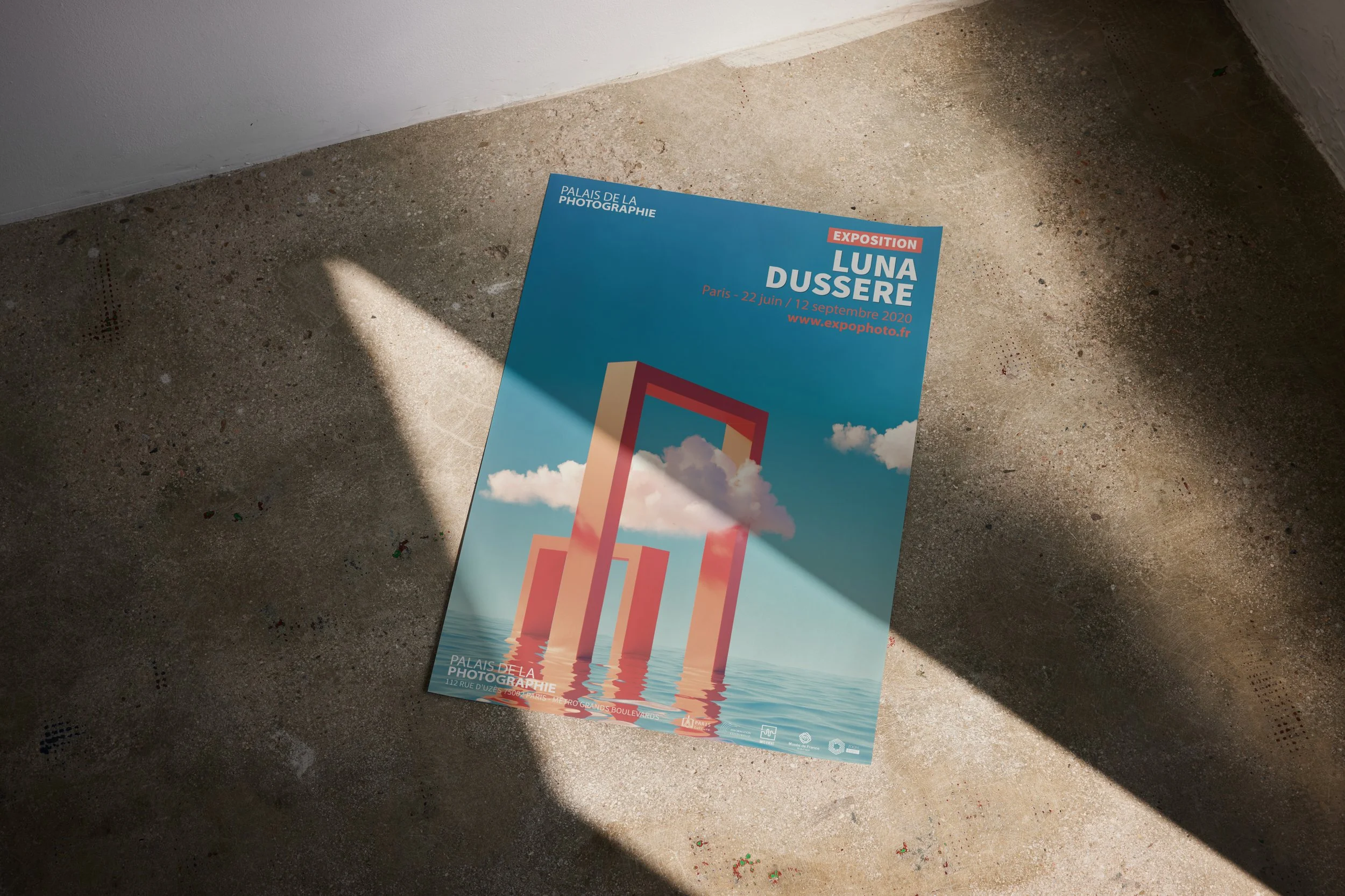 Affiche d'une exposition au Palais de la Photographie à Paris, du 22 juin au 12 septembre 2020, avec une image de structures rectangulaires réfléchies dans l'eau et un ciel bleu avec des nuages.