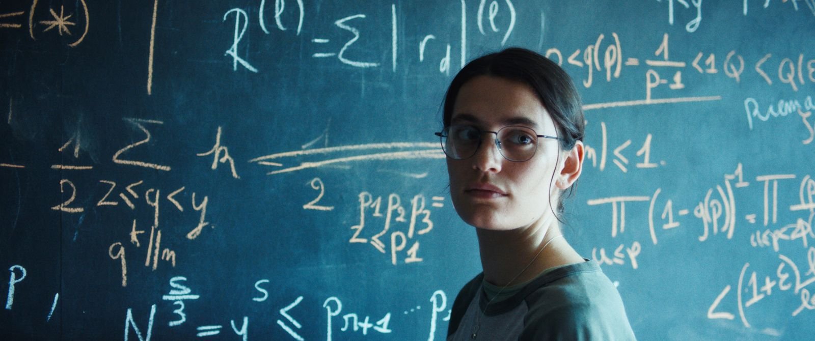 Une jeune femme portant des lunettes regarde vers l'objectif, debout devant un tableau noir rempli d'équations mathématiques en craie.