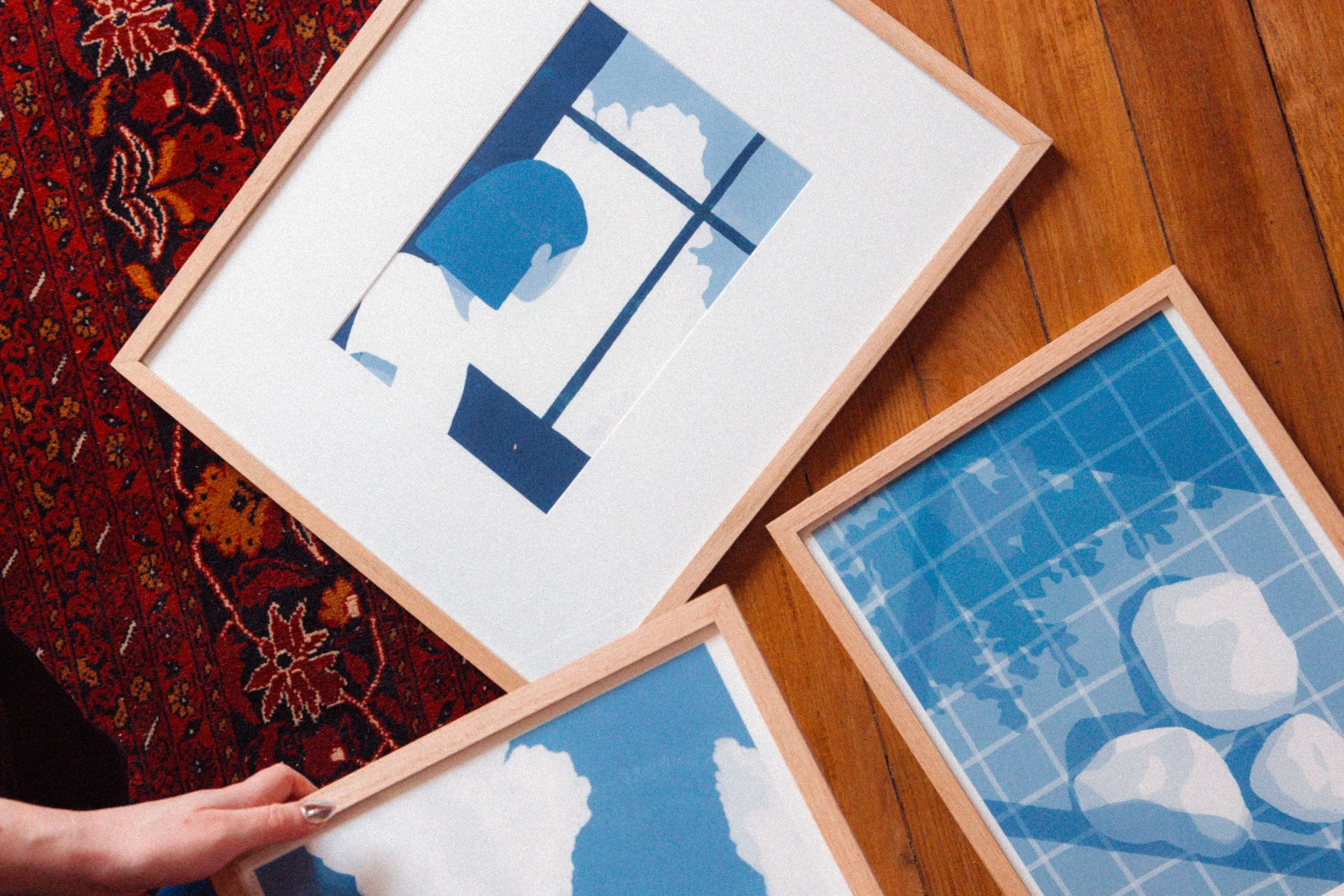 Quatre œuvres d'art modernes avec des tons bleus, encadrées en bois, posées sur un sol en bois et une tapis rouge.