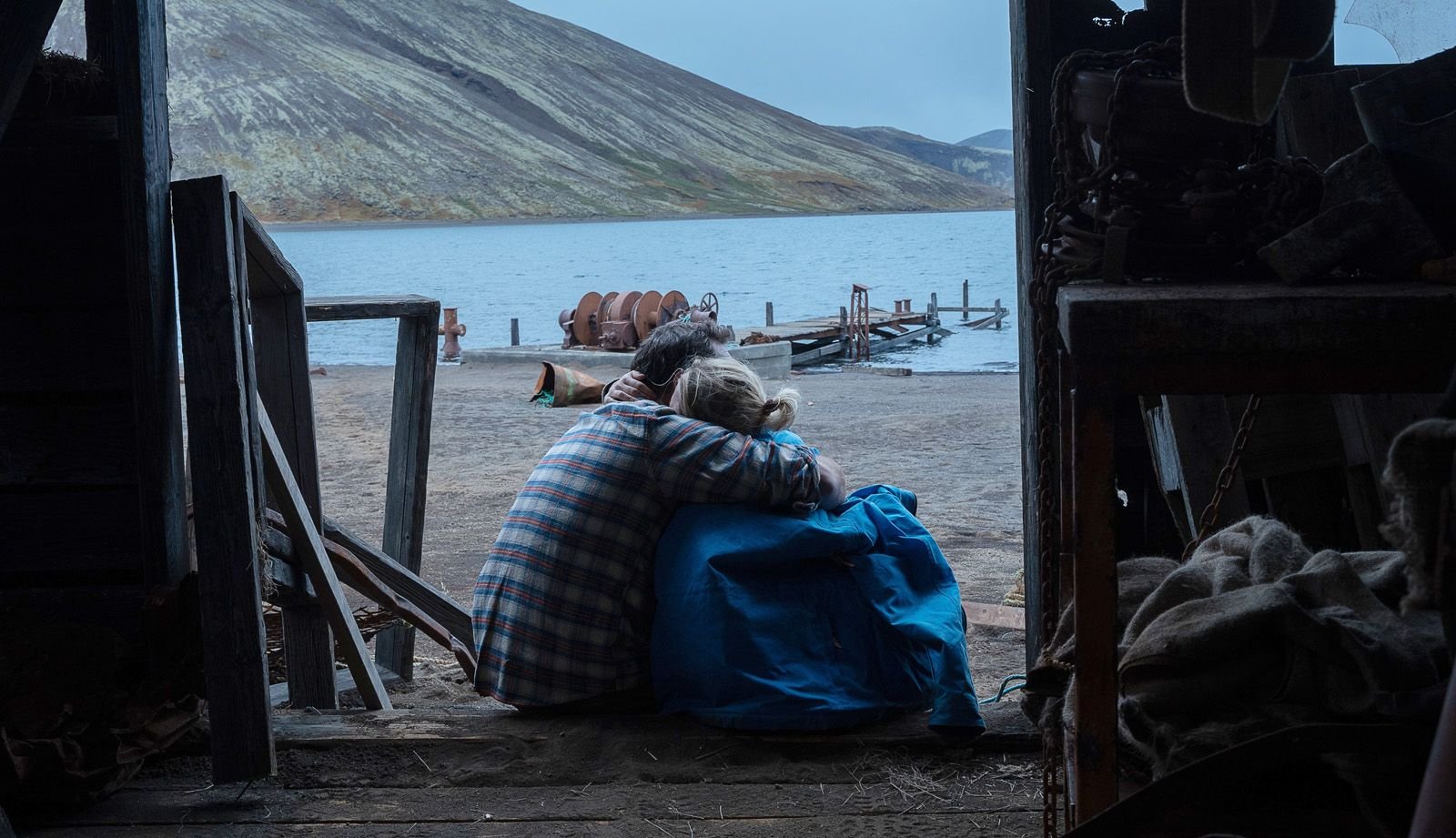 Deux personnes assises côte à côte, regardant un lac depuis l'entrée d'une cabane en bois, entouré de montagnes.