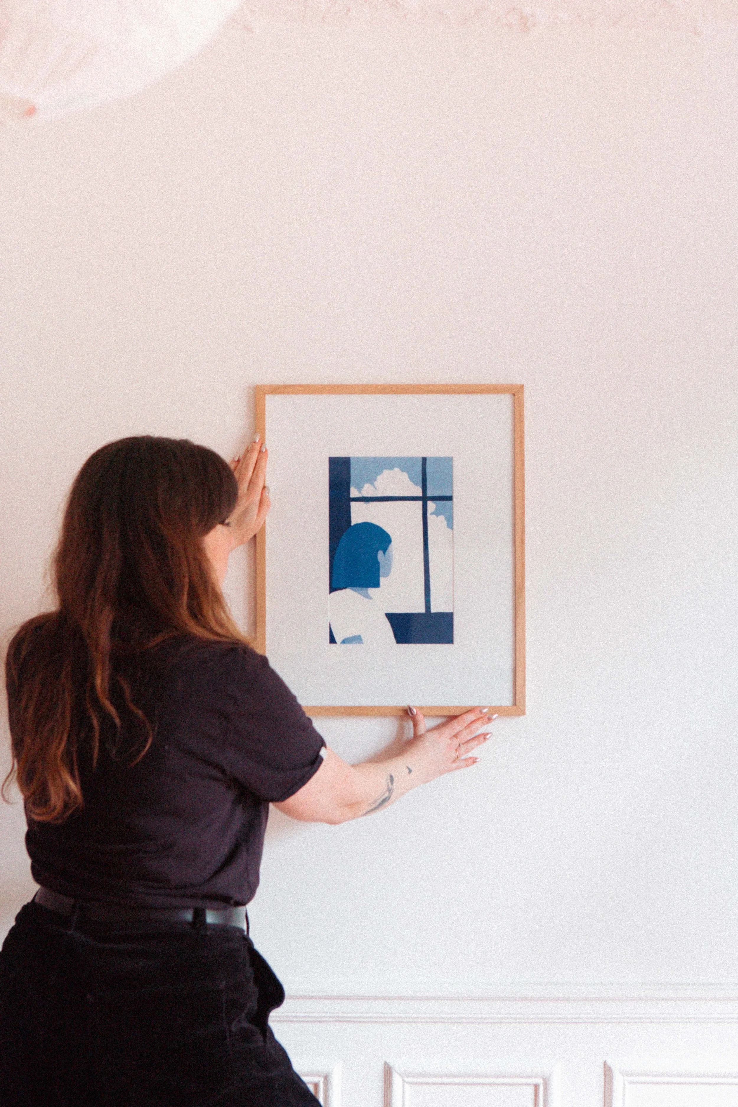 Une femme accroche un tableau avec une illustration de profil d'une personne regardant par une fenêtre, en intérieur.