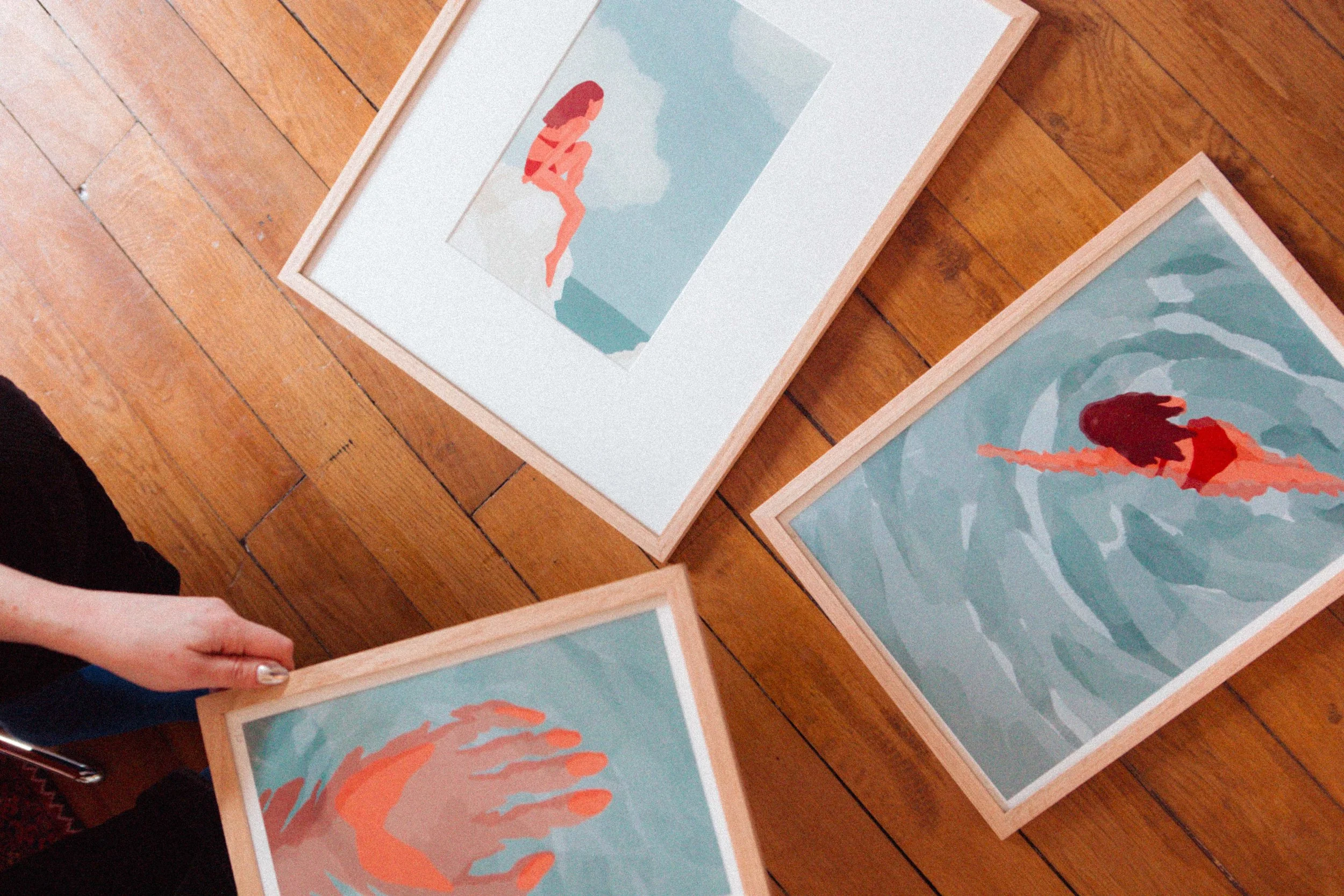 Trois tableaux avec des illustrations de femmes dans différentes poses sur des fonds aquatiques, disposés sur un sol en bois.