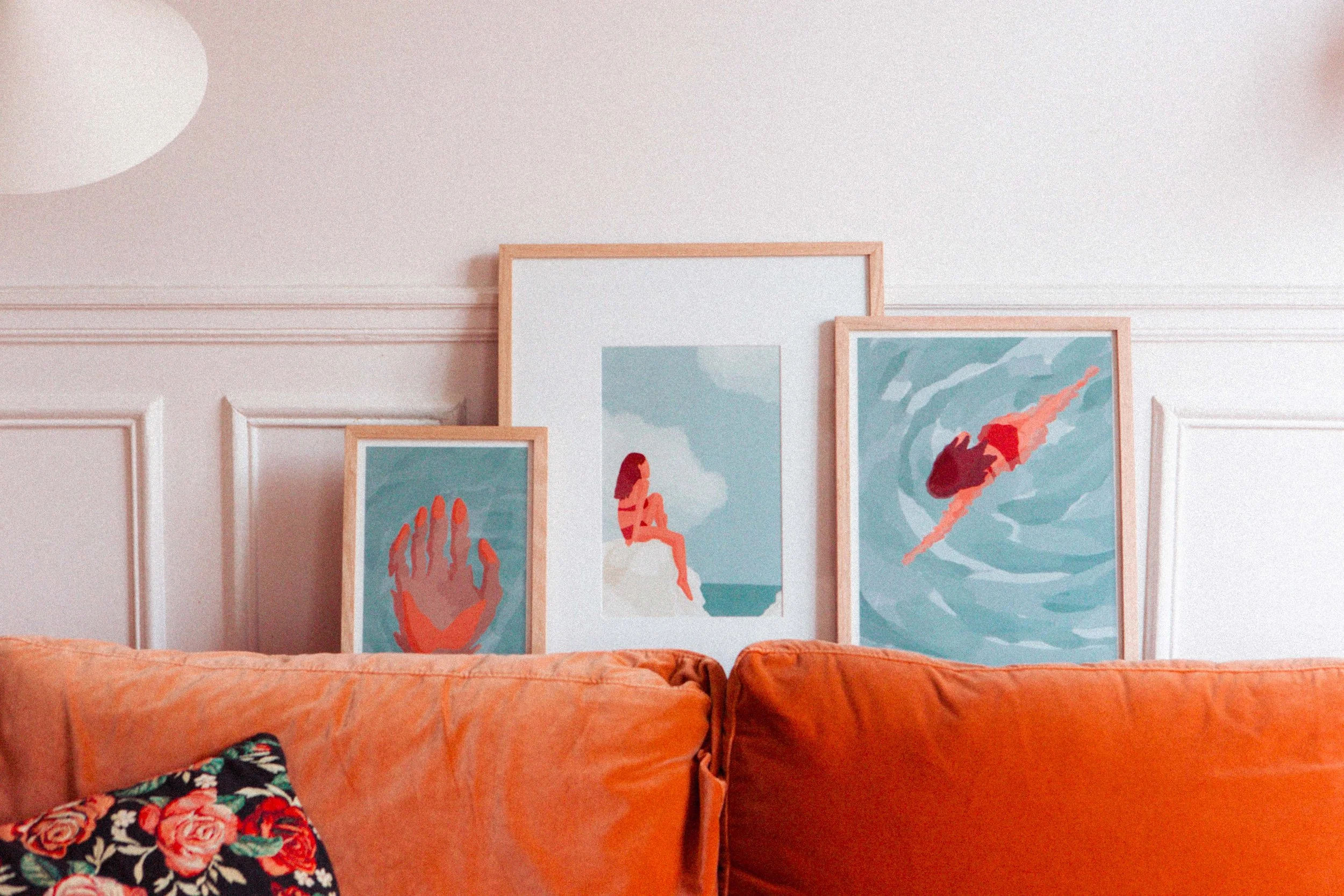 Trois tableaux de style graphique, représentant une main, une femme assise et un nageur dans des tons bleus et rouges, reposant sur un canapé orange.