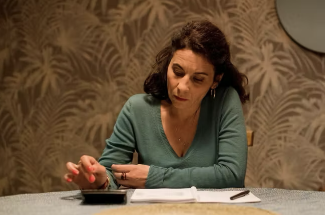 Femme assise à une table, regardant un téléphone portable, avec un carnet et un stylo devant elle, fond avec motif de feuilles.