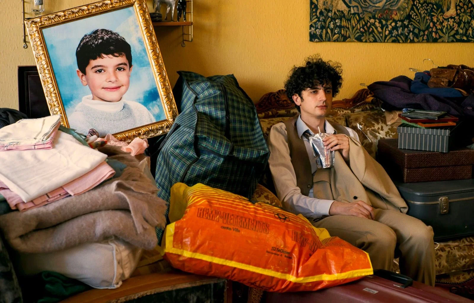 Un jeune homme à cheveux bouclés assis sur un canapé en train de boire dans un paquet de jus. Sur le canapé, il y a des piles de vêtements, des sacs et une boîte à linge. À côté de lui, une grande photo encadrée d'un enfant souriant, ainsi que des valises et des sacs sur une table et en arrière-plan. La pièce est décorée avec un mur jaune, une tenture et des étagères.