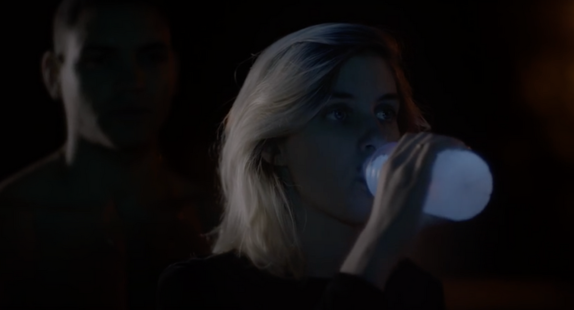 Une femme blonde boit dans une bouteille dans un environnement sombre, avec une autre personne floue en arrière-plan.