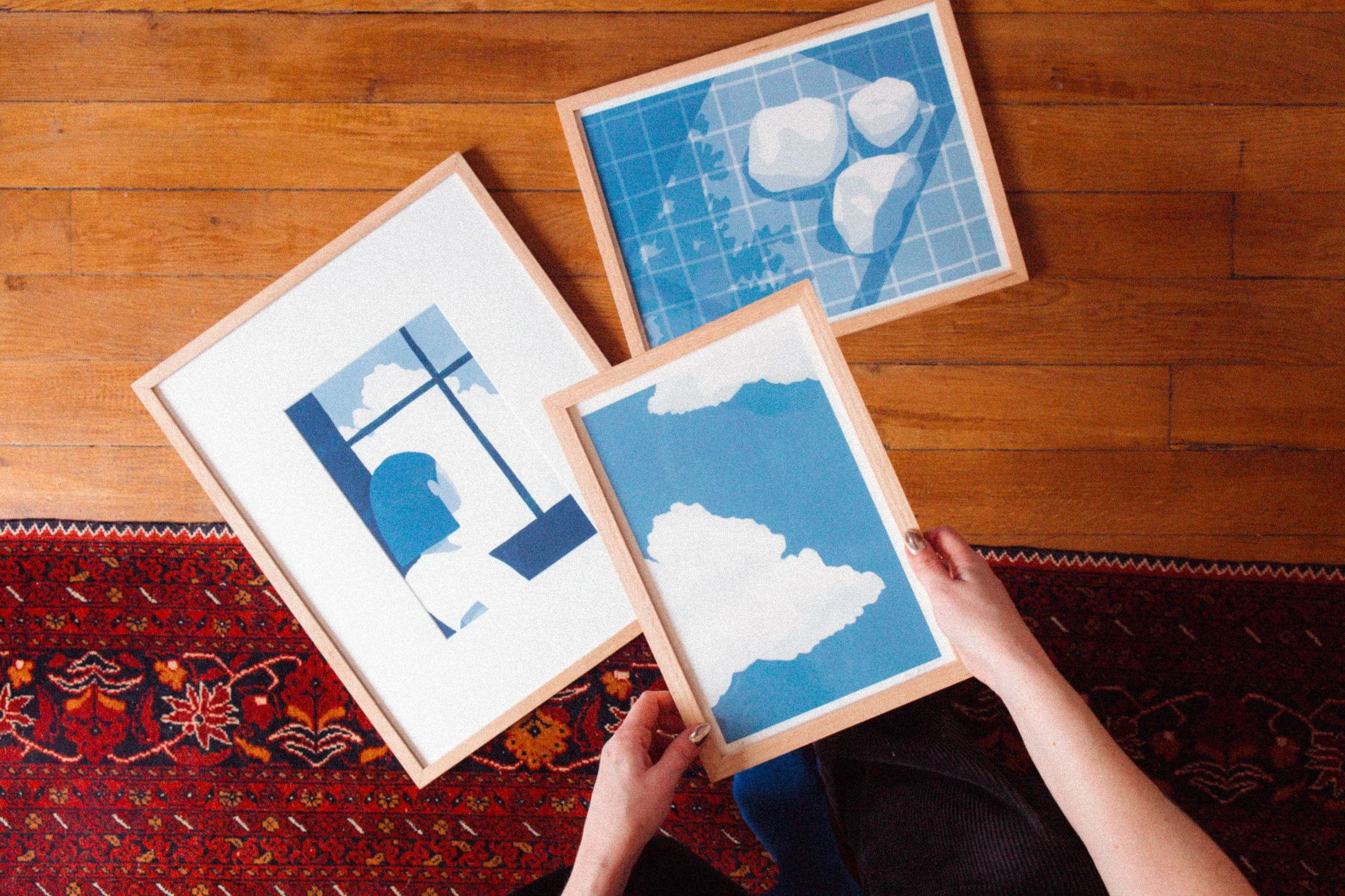 Personne tenant trois tableaux, un avec un paysage de montagne, un avec des nuages, un avec des rochers, posé sur un tapis rouge et un sol en bois.