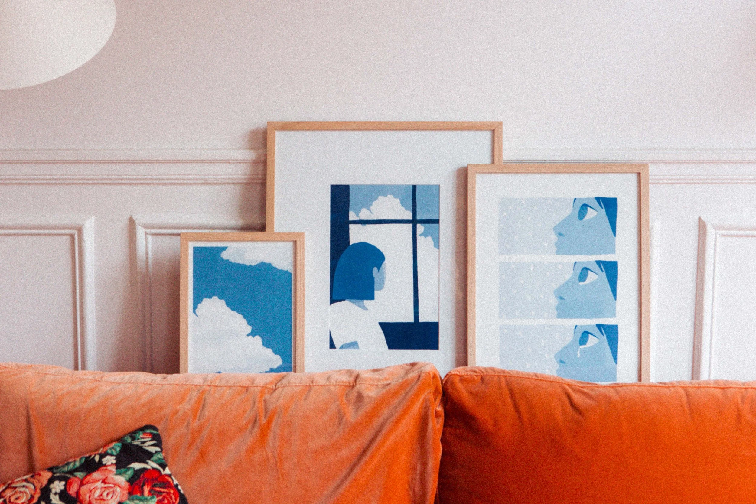 Quatre cadres avec illustrations bleues sont posés derrière un canapé orange en velours. La première illustration montre un ciel avec des nuages, la deuxième une femme regardant par une fenêtre, et la troisième une femme en profil dans plusieurs expressions.