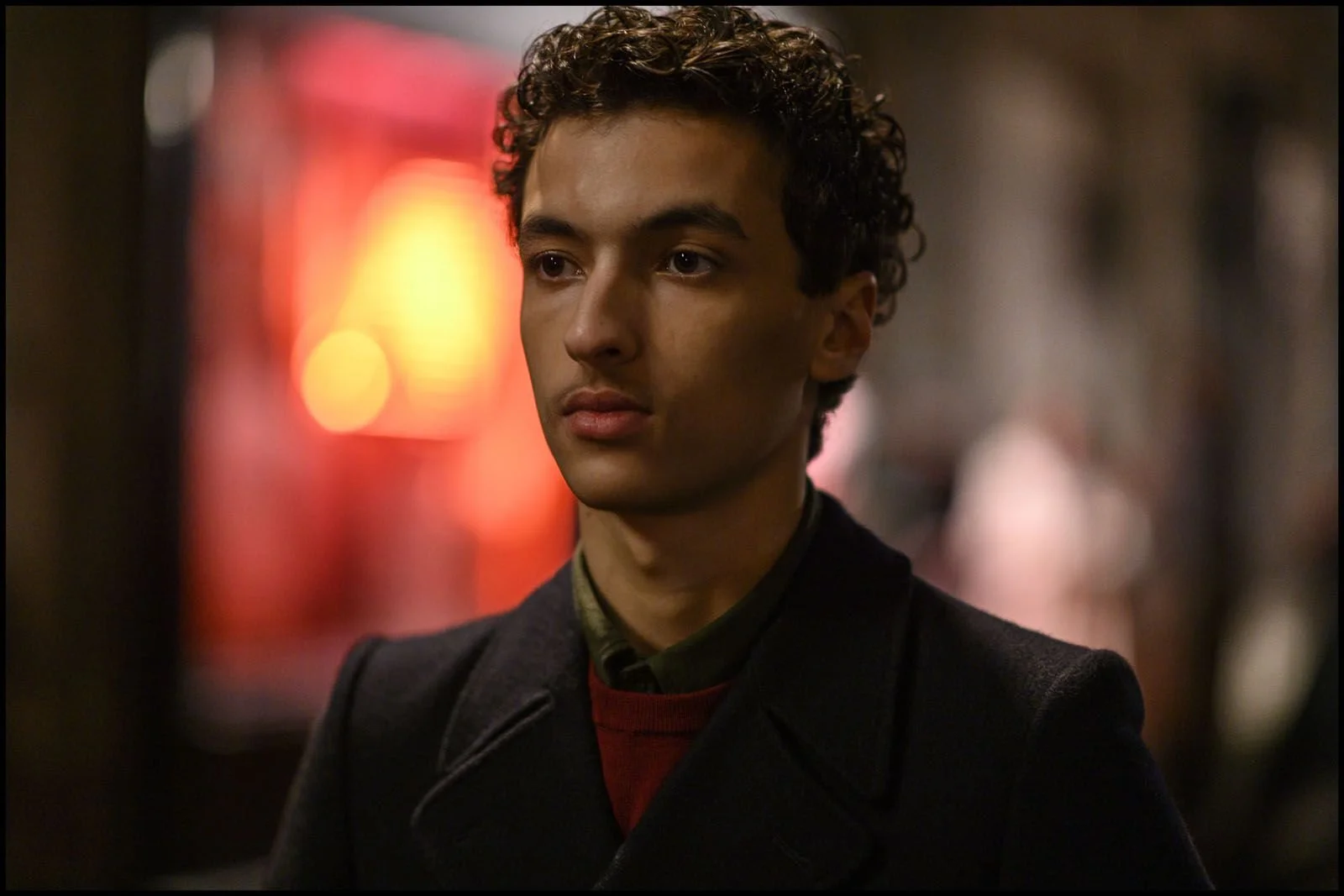 Jeune homme avec des cheveux bouclés foncés, portant un manteau noir, dans une ambiance urbaine nocturne avec des lumières floues en arrière-plan.