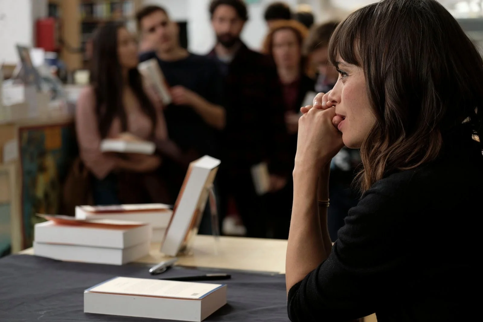 Une femme assise à une table, semble en attente, avec plusieurs livres devant elle, et un groupe de personnes flou en arrière-plan dans un espace intérieur.