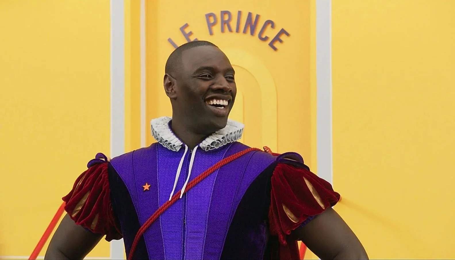 Une personne souriante déguisée en prince avec un costume coloré, devant un mur jaune avec le texte 'Le Prince'.