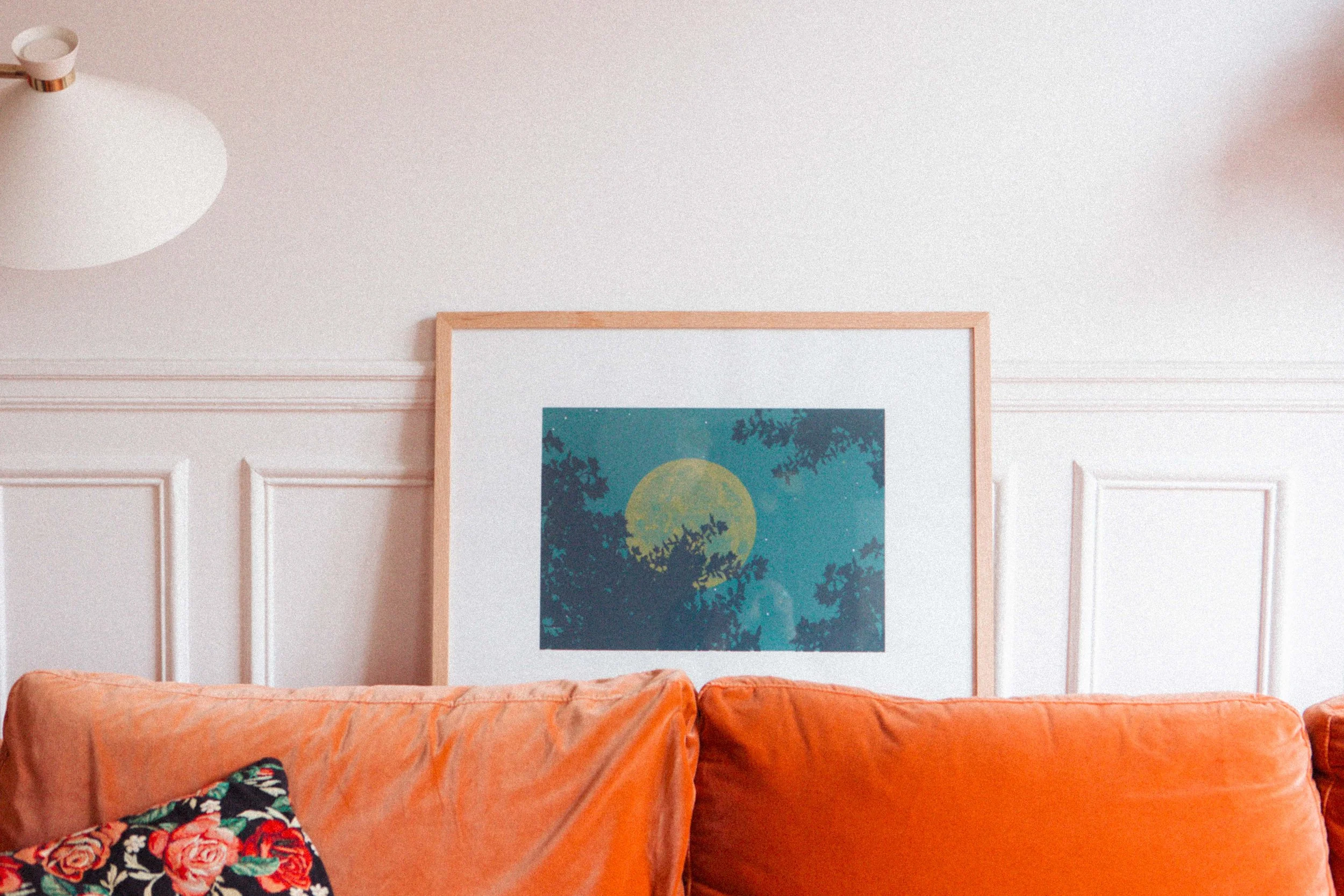 Tableau représentant une lune pleine derrière des branches d'arbres dans une pièce avec un canapé orange et une lampe blanche au plafond.