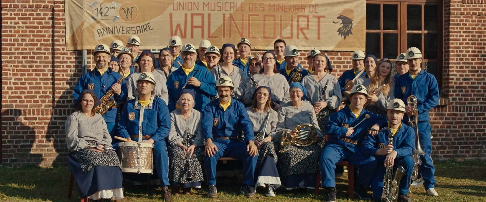Photo d'un groupe de musiciens habillés en costumes bleus et gris, portant des casques avec lampe, posant devant un mur en briques avec une bannière annonçant le 142ème anniversaire de l'Union musicale des Mineurs de Walldorf.