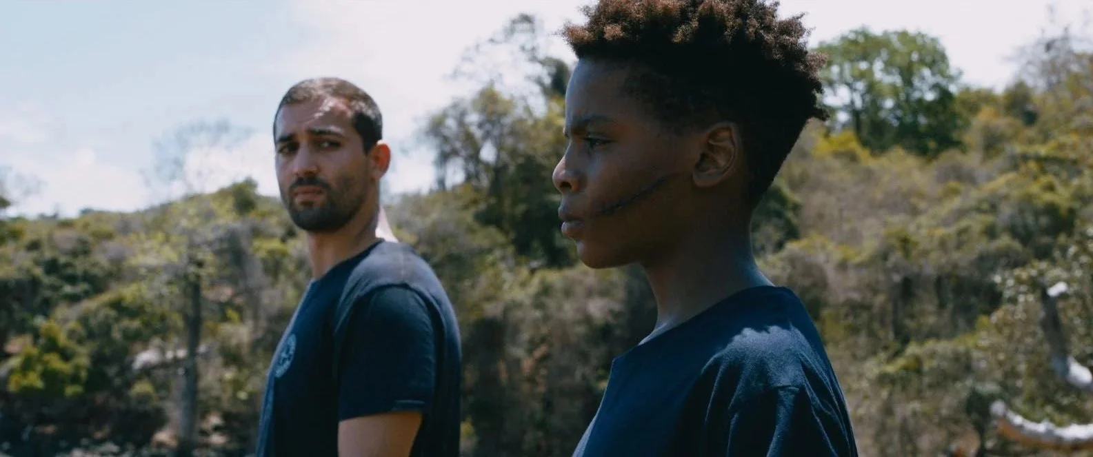 Deux jeunes hommes regardent dans des directions opposées, l'un avec une expression préoccupée, l'autre avec un visage sérieux, dans un environnement extérieur avec des arbres en arrière-plan.