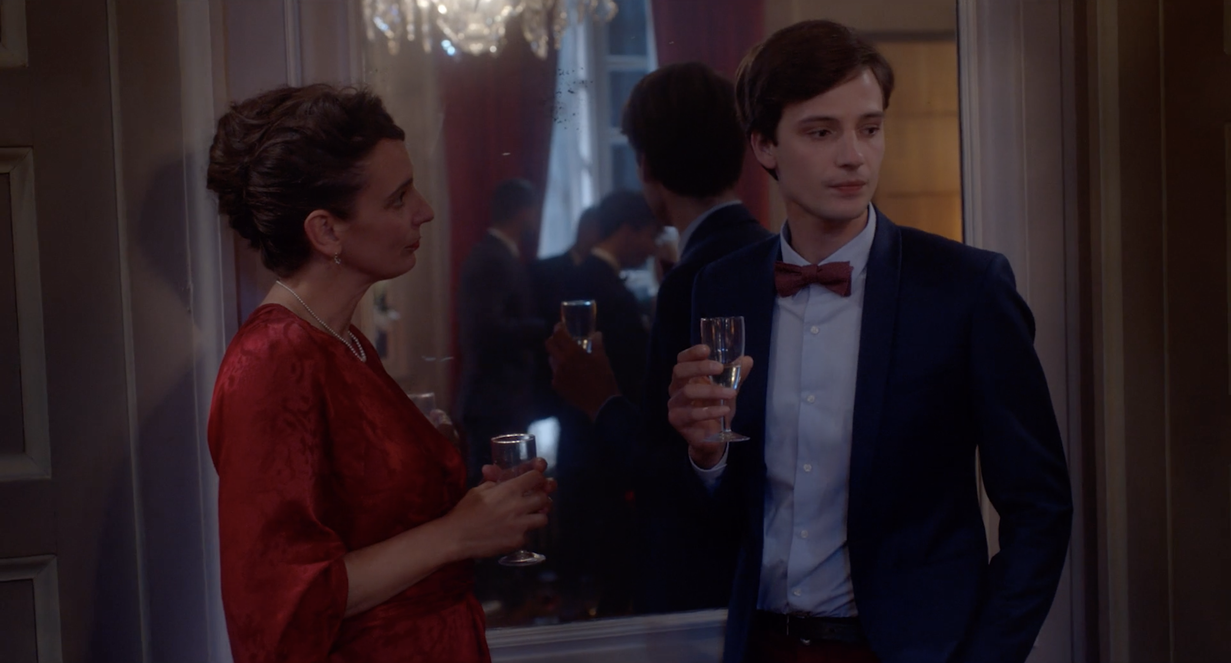 Une femme et un jeune homme en costume discutent lors d'une fête ou d'un événement social, tenant des flûtes de champagne.
