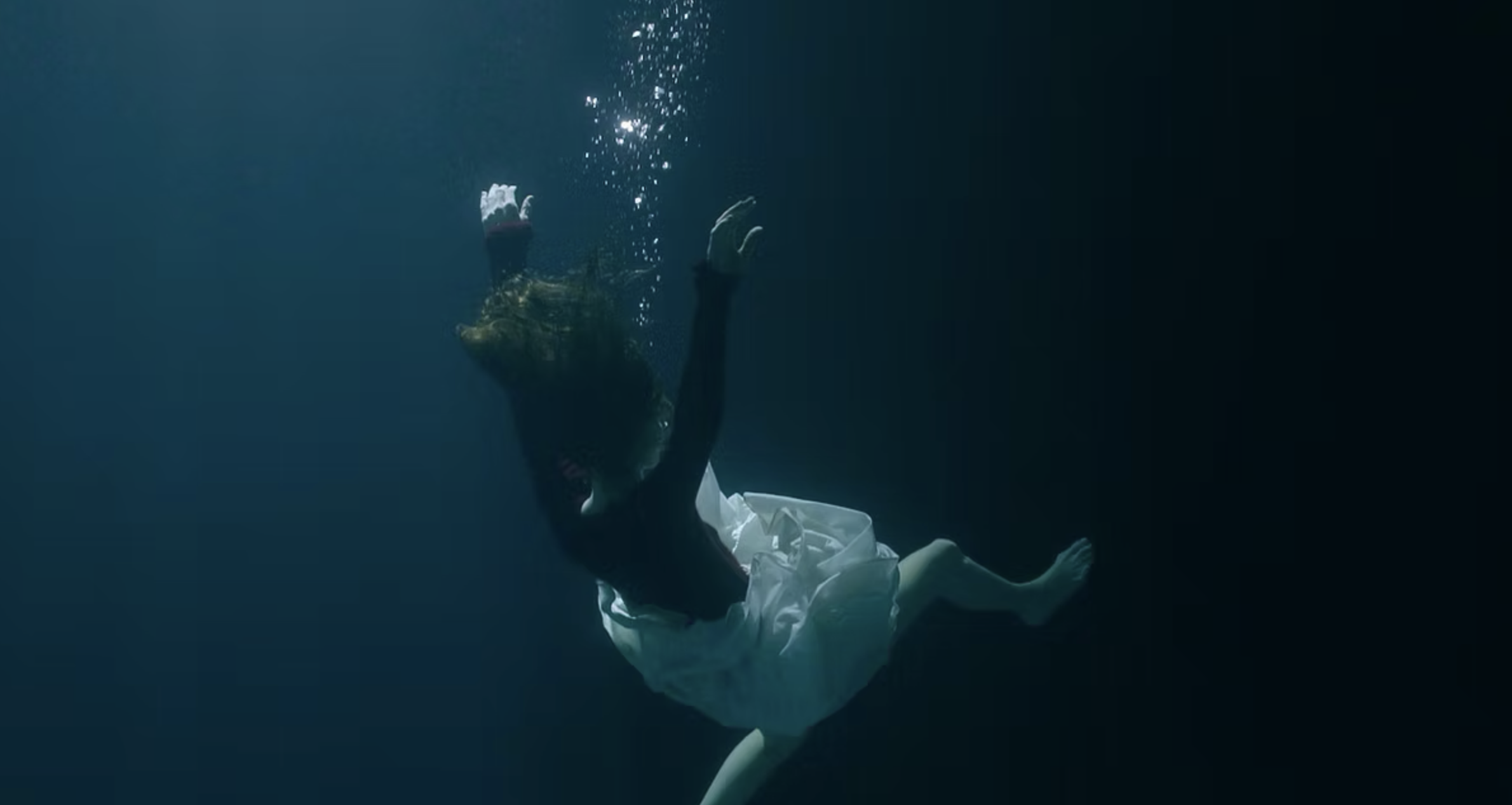 Une personne en robe blanche nage sous l'eau, avec des bulles d'air s'échappant de sa bouche ou de son nez.