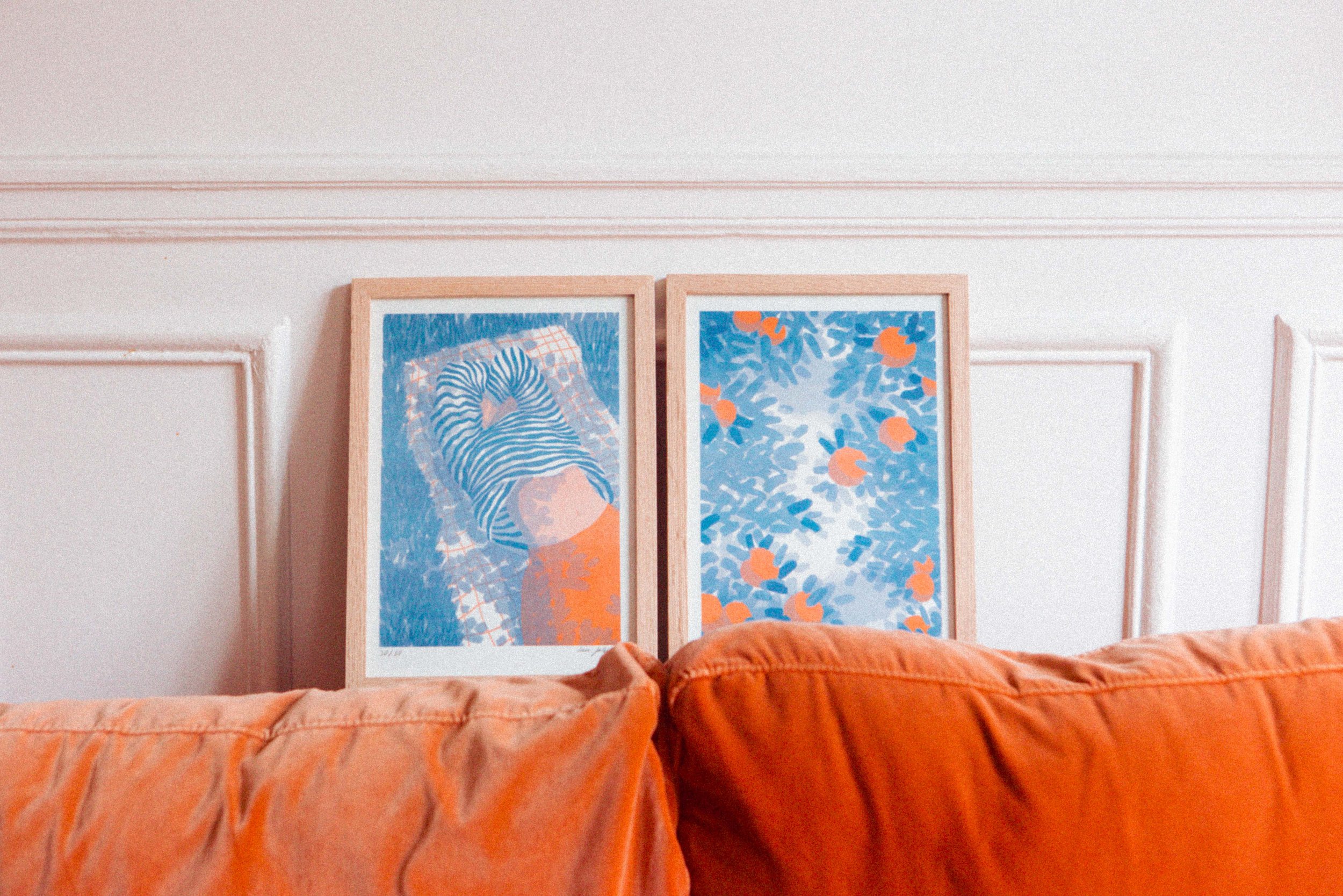 Deux tableaux encadrés reposant contre un mur blanc au-dessus d'un canapé orange, un dos d'une personne avec un t-shirt à rayures est visible sur la gauche.