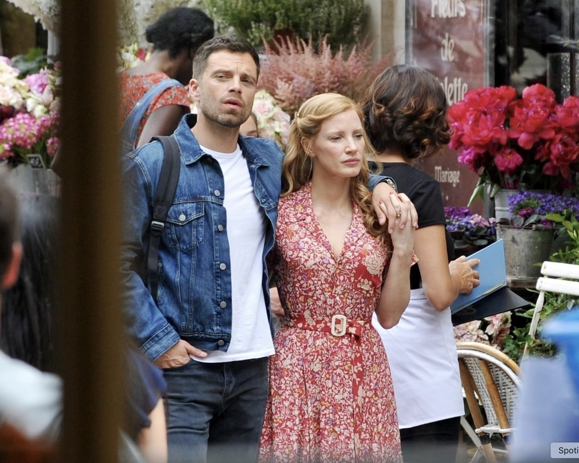 Deux personnes, un homme et une femme, se tiennent côte à côte dans une rue bordée de fleurs, manifestant de la tristesse ou de la prière par leurs expressions et leur posture, lors d'une journée en plein air.