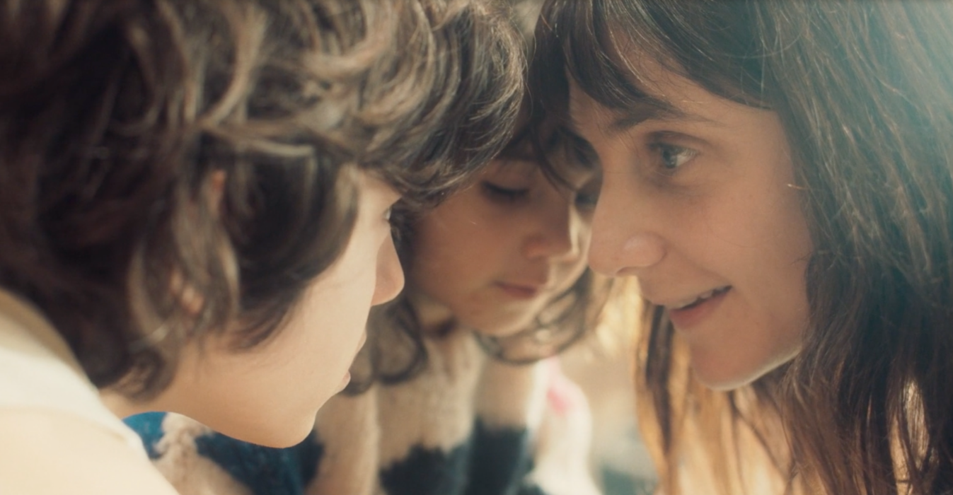 Trois personnes, deux enfants et une femme, sont très proches, face à face, avec leurs visages presque collés, partageant un moment intime et concentré.