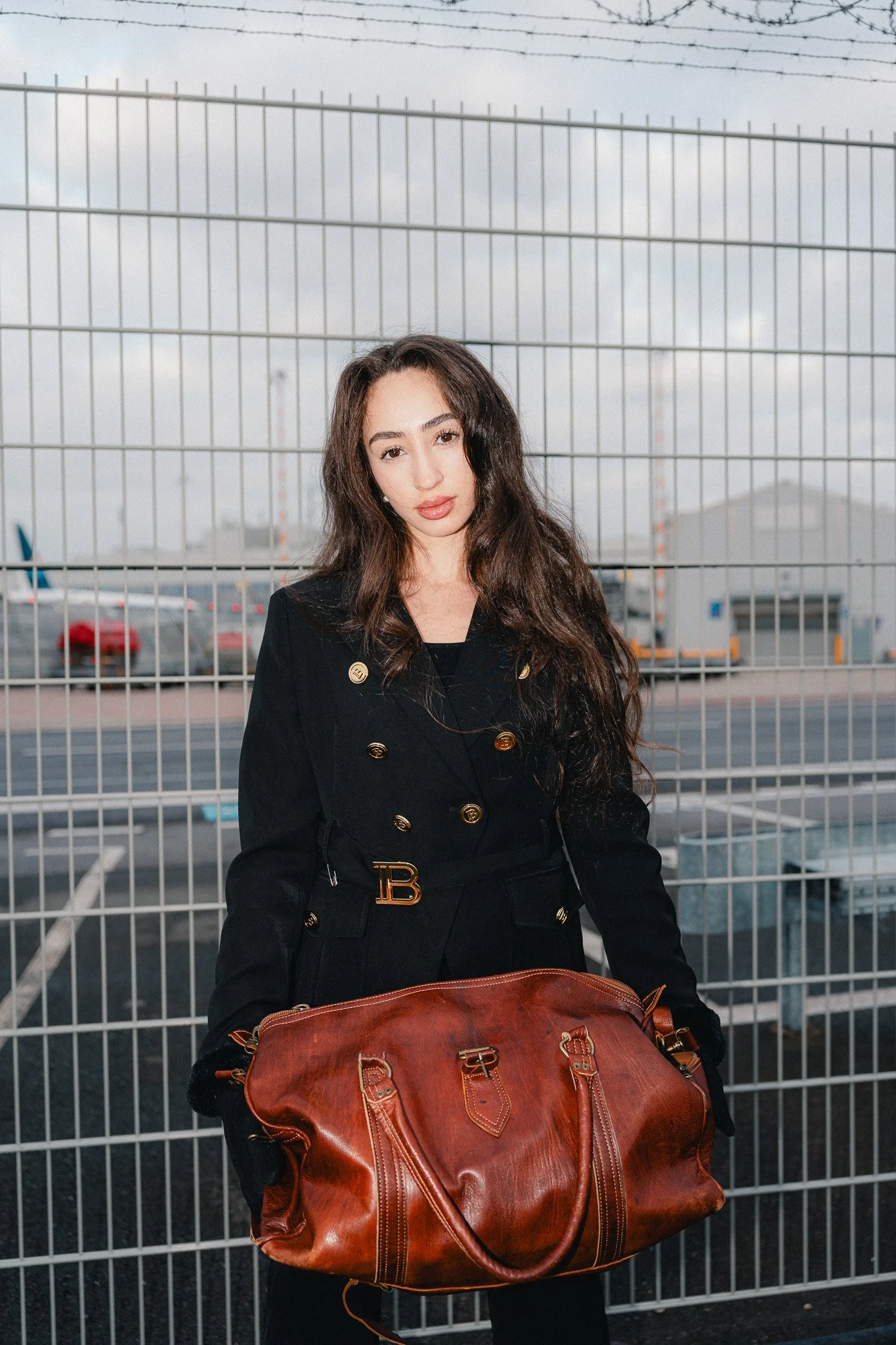 Eine junge Frau hält eine große braune Ledertasche vor einem Flughafen-Gitterzaun. Im Hintergrund sind Flugzeuge und Flugzeugtarmac sichtbar, bei bewölktem Himmel.
