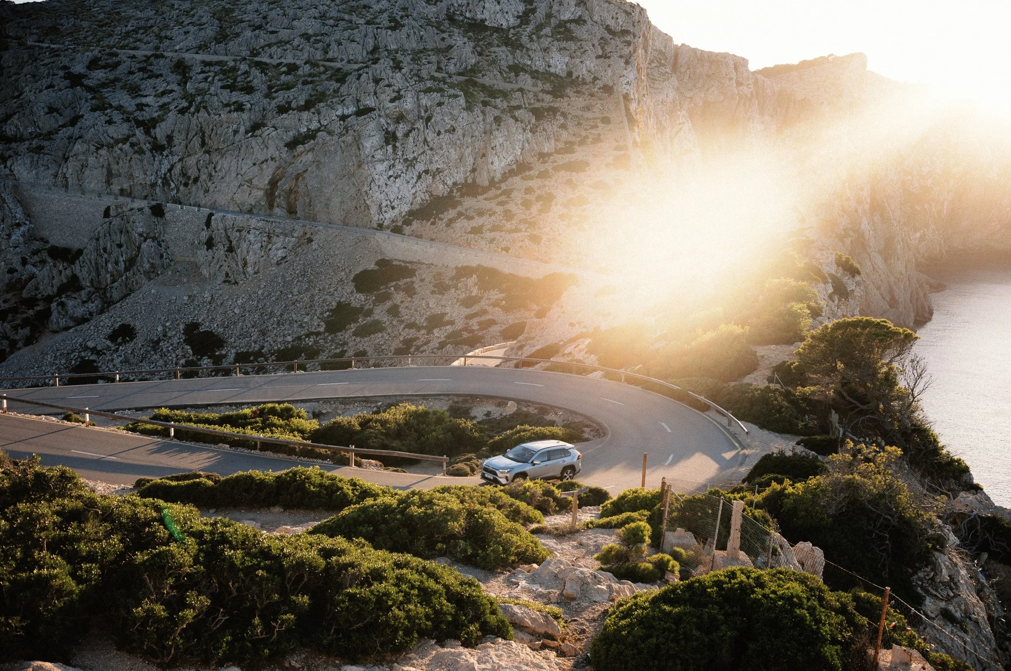 Kurvige Autobahn führt an einer Steilküste entlang, Sonnenlicht strahlt durch die Bäume, eine weiße SUV ist unterwegs.
