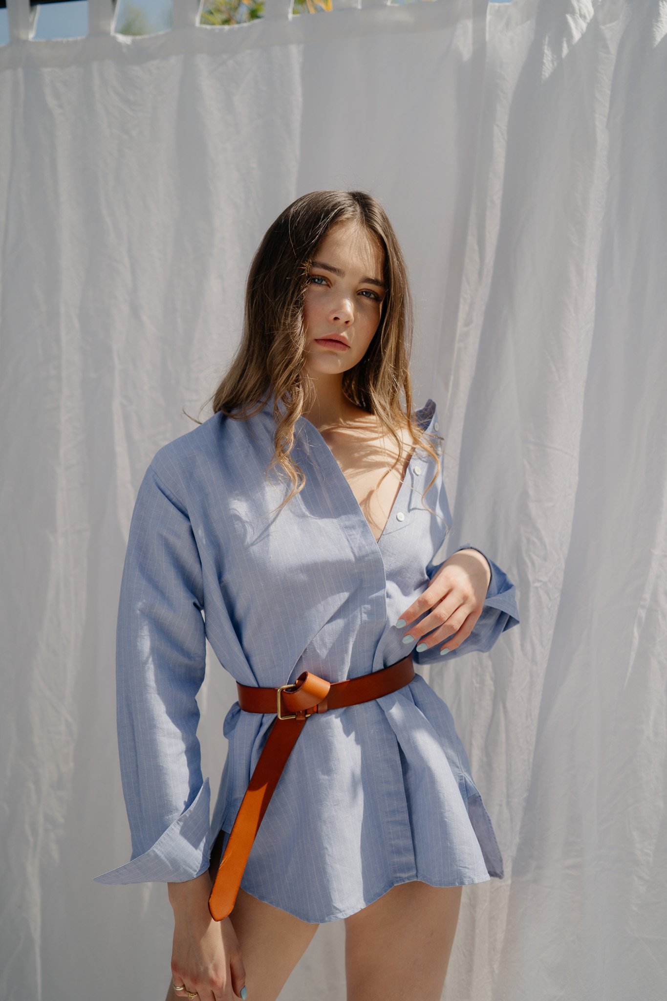 Junge Frau in blauer Hemdbluse mit braunem Gürtel, steht vor einer weißen Stoffwand im Sonnenlicht.