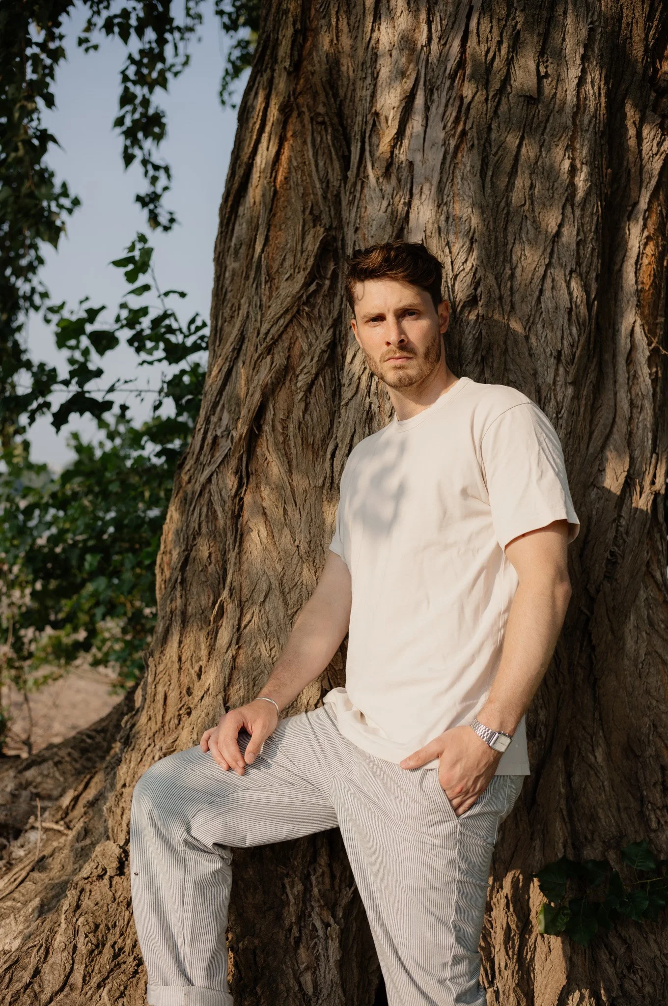 Ein junger Mann lehnt an einem großen Baum im Freien, trägt ein weißes T-Shirt und helle, gestreifte Hosen, schaut ernst in die Kamera, mit einer Armbanduhr am Handgelenk.