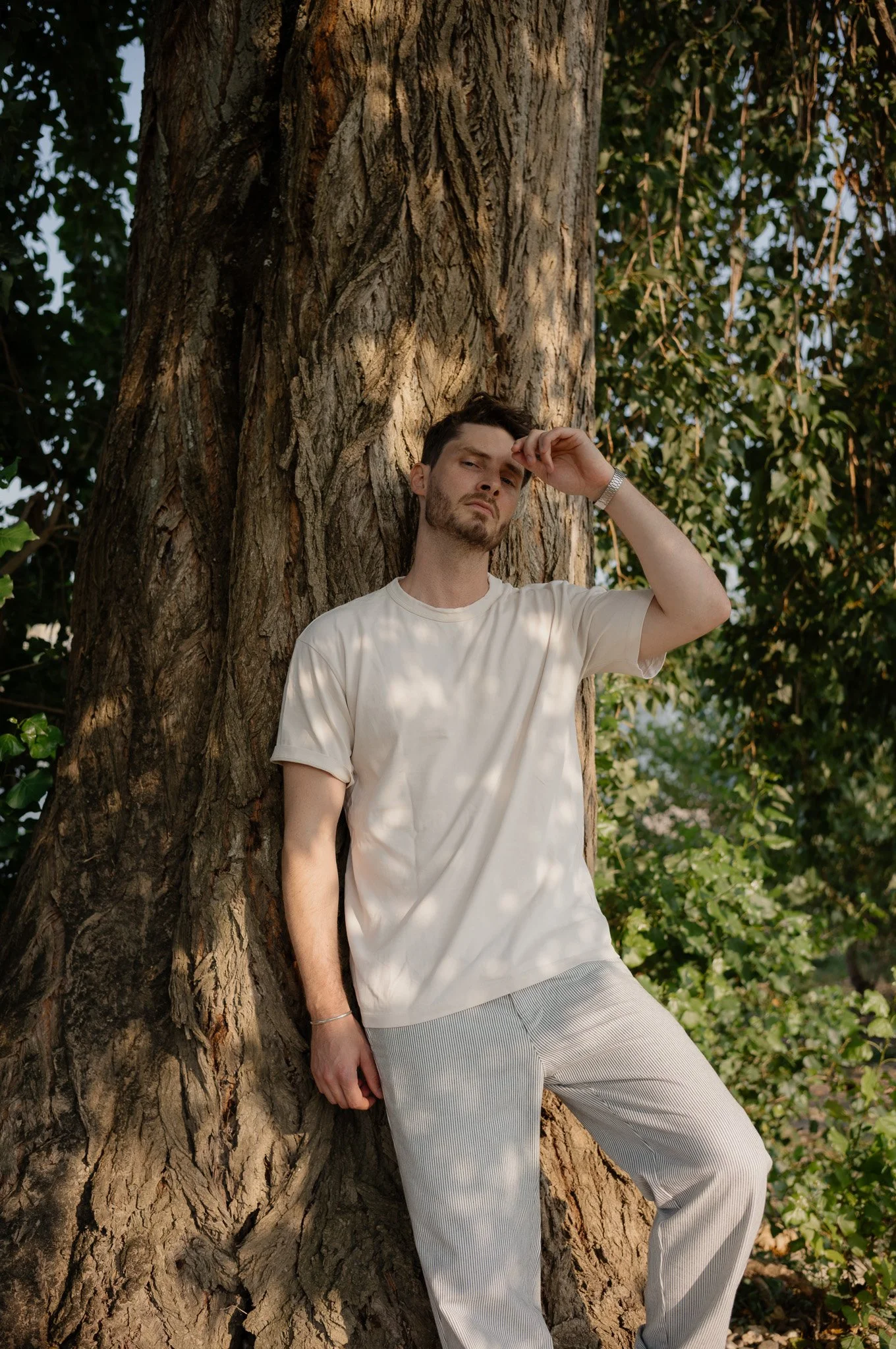 Junger Mann lehnt an einem Baum im Grünen, trägt ein weißes T-Shirt und hellgraue Hosen, schaut ernst durch die Haare gehalten.