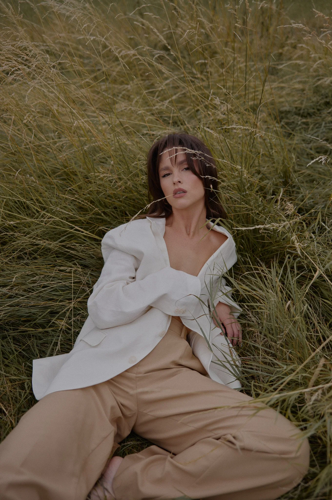 Frau liegt in hohem Gras, trägt beige Hosen und eine weiße Jacke, die halb offen ist, mit zerzaustem Haar und einem entspannten Gesichtsausdruck.