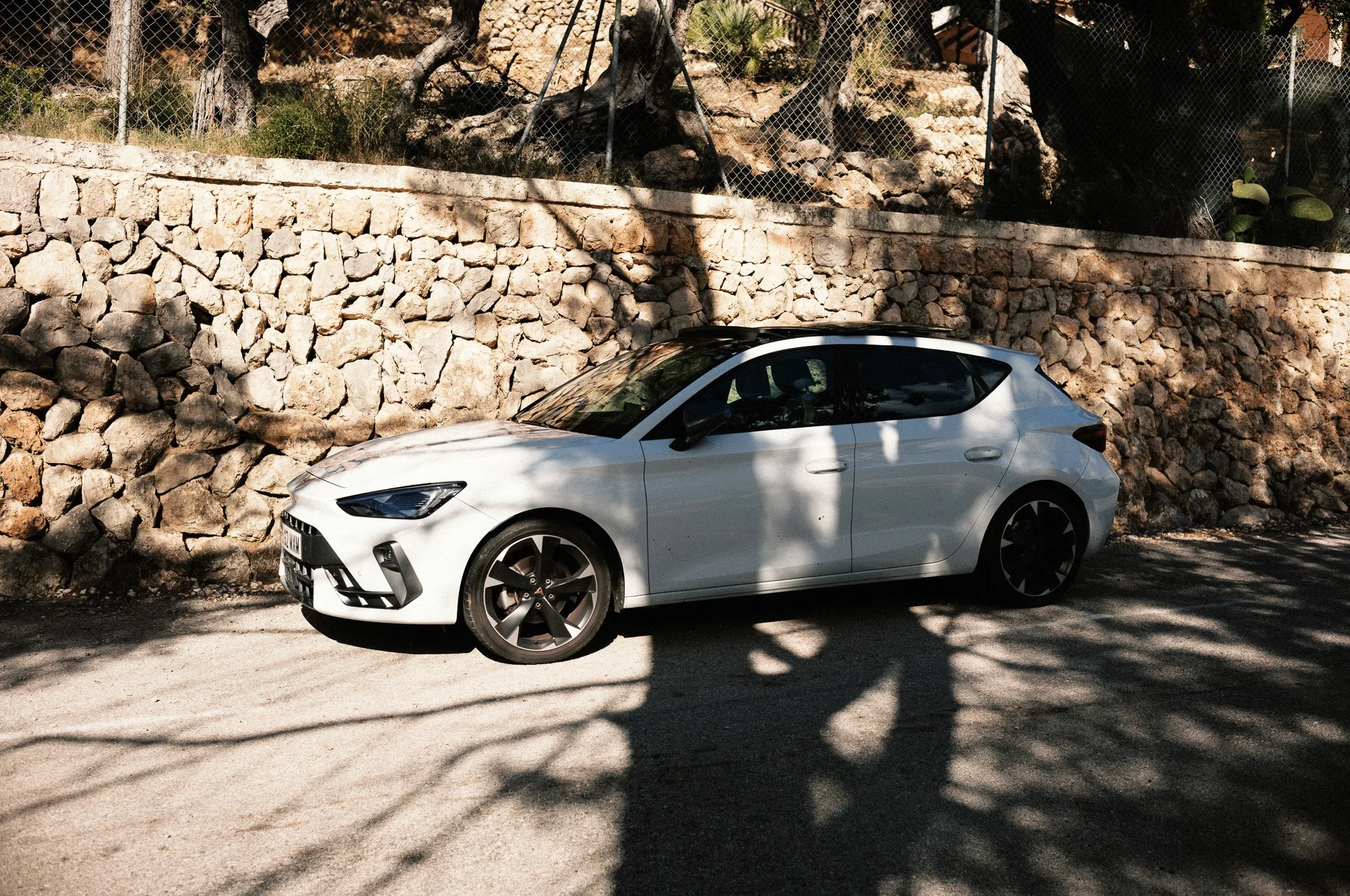 Ein weißes Auto steht am Straßenrand, im Schatten eines Baumes, vor einer Natursteinmauer mit Bäumen dahinter.