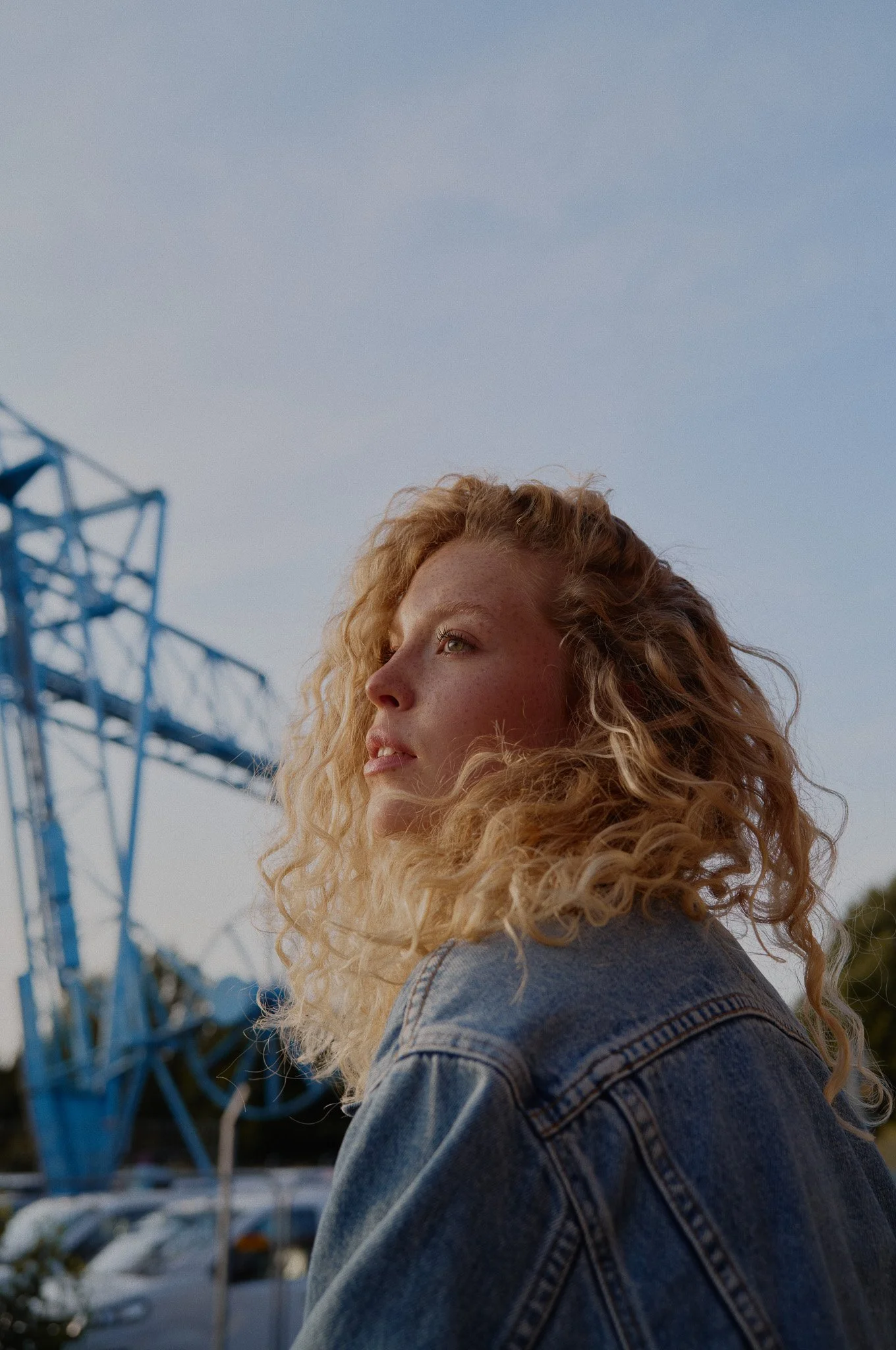 Junge Frau mit lockigem Haar trägt eine Jeansjacke und schaut nachdenklich in die Ferne bei Sonnenuntergang, im Hintergrund eine Industriekonstruktion und der Himmel.