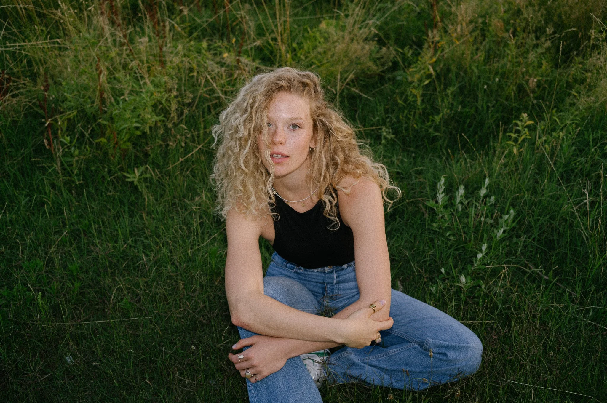 Junge Frau mit lockigem blondem Haar, schwarzen Top und Jeans sitzt auf grünem Gras in einer Wiese.