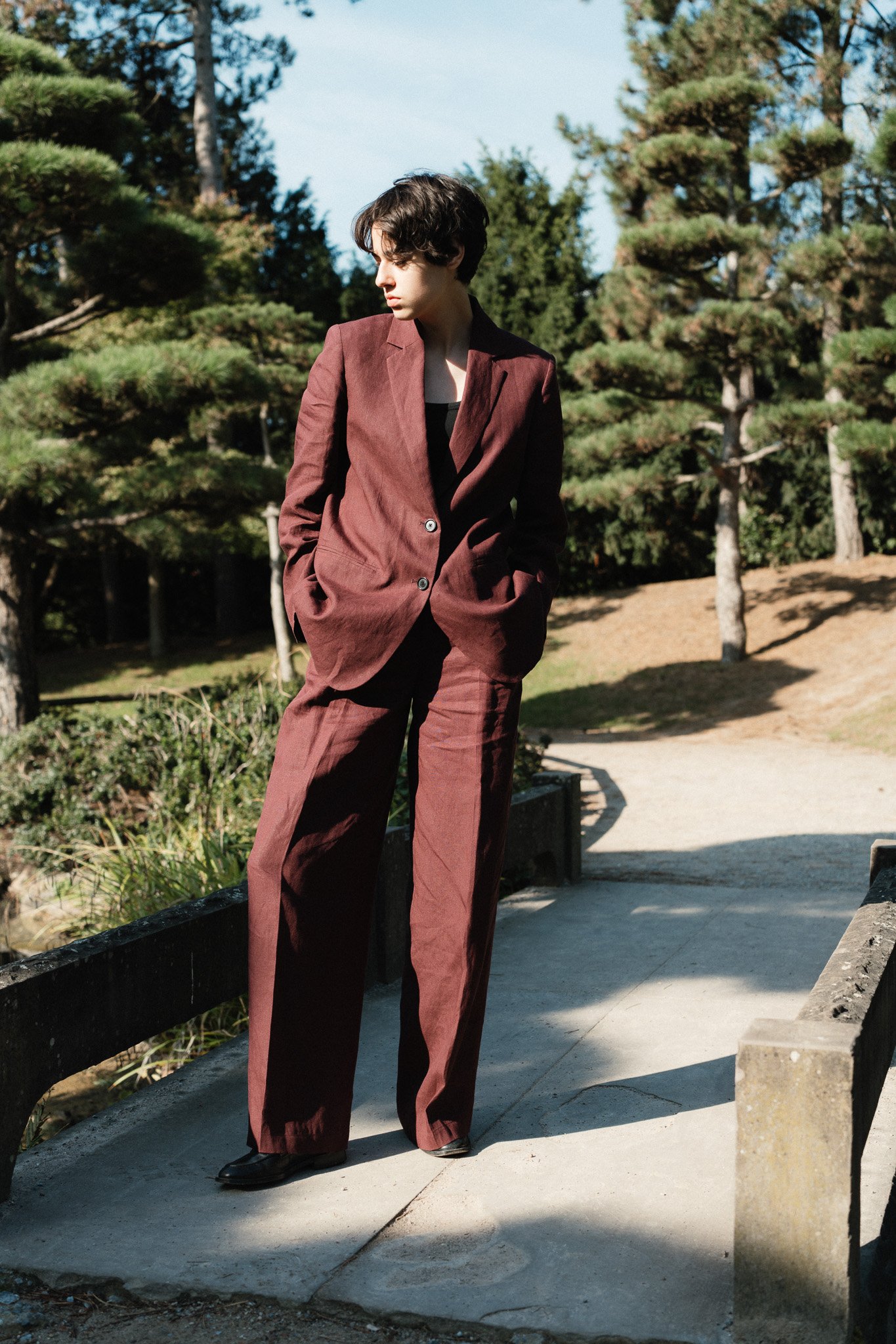 Junge Frau in braunem Anzug steht an einer Brücke im Park, während die Sonne scheint.
