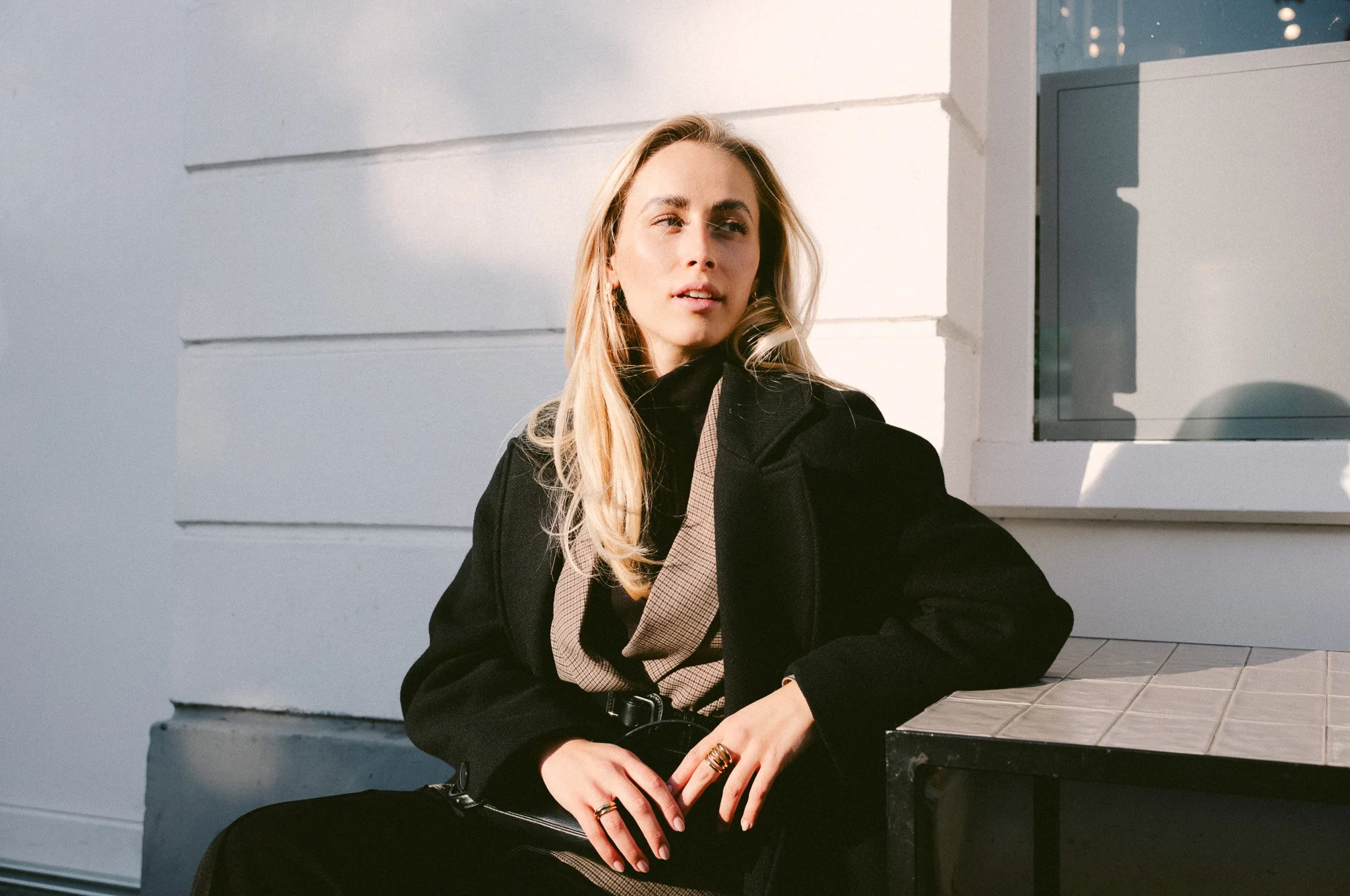 Eine blonde Frau mit schwarzer Jacke sitzt an einem Tisch vor einer weißen Wand.