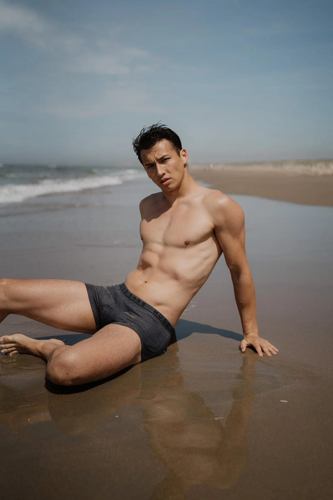 Ein junger Mann sitzt am Strand im Sand, trägt nur Unterwäsche, schaut direkt in die Kamera, bei sonnigem Himmel und Wellen im Hintergrund.