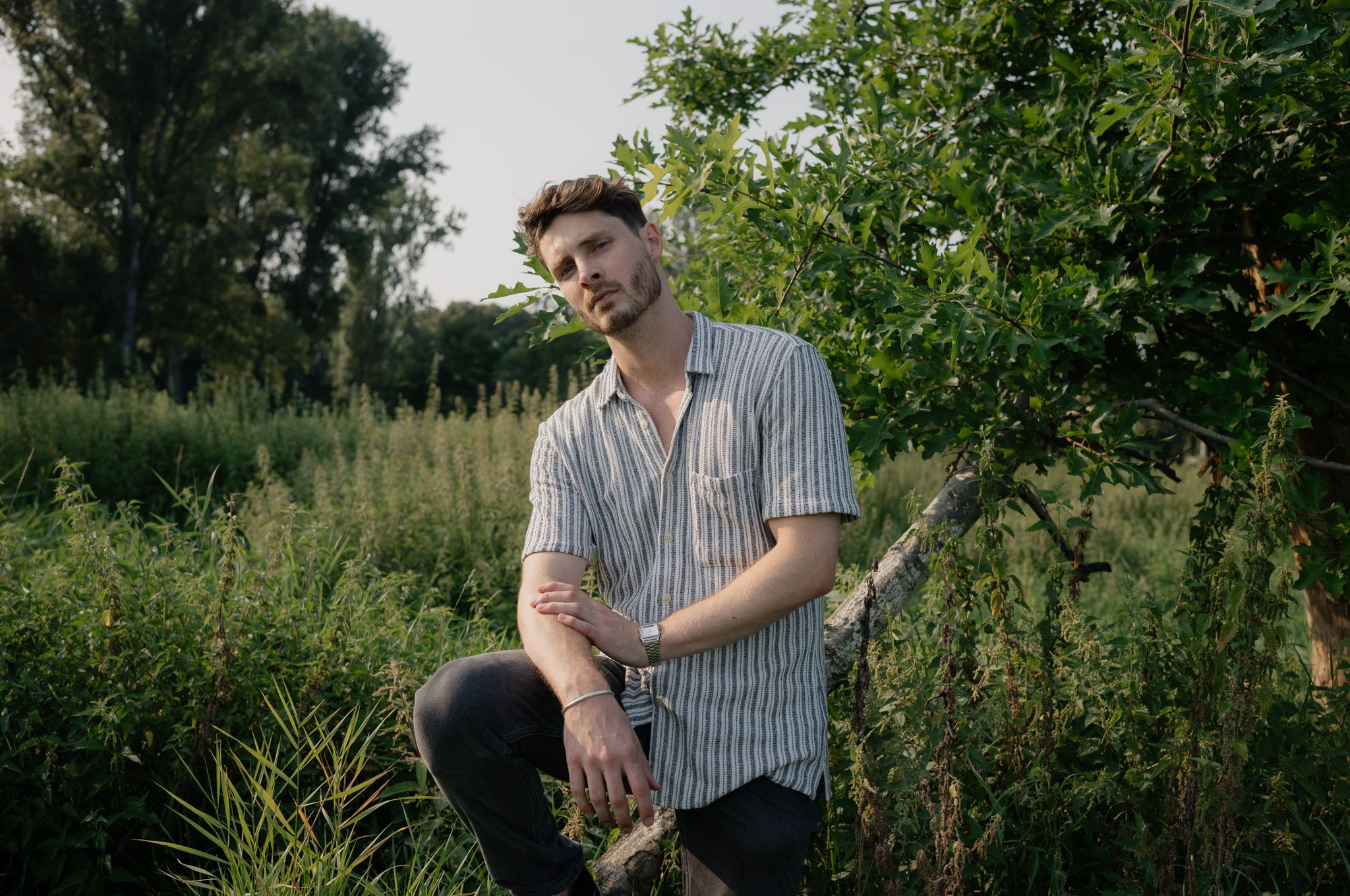 Ein junger Mann mit kurzen braunen Haaren und Bart, trägt eine grau-weiß gestreifte Hemd, sitzt an einem Baum im Grünen, umgeben von Pflanzen und Bäumen, bei Sonnenuntergang.