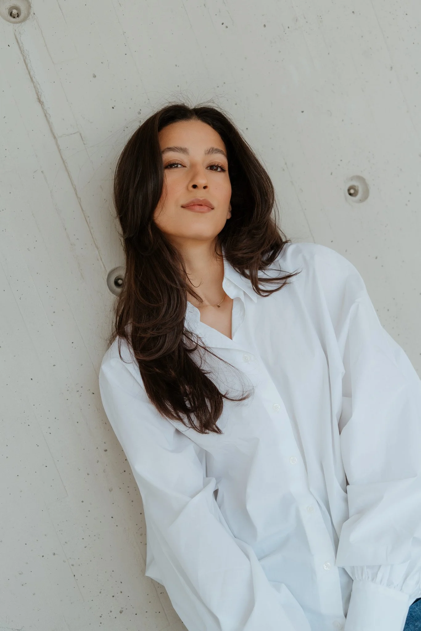 Junge Frau in weißem Hemd lehnt an einer Betonwand, Blick in die Kamera, lockere braune Haare.