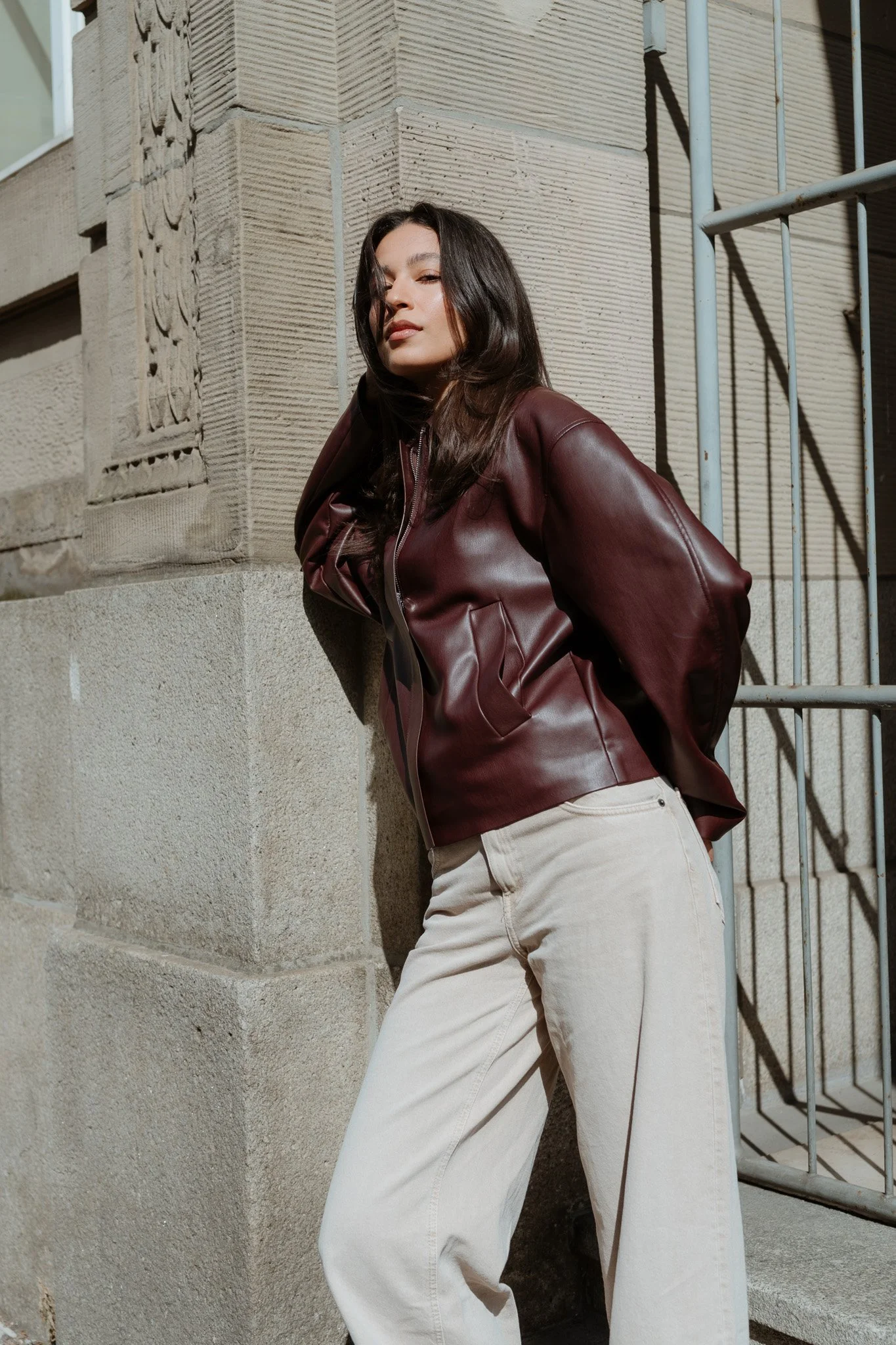 Junge Frau lehnt an einer Steinmauer, trägt eine braune Lederjacke und hellbeige Hosen, Sonne scheint auf sie.