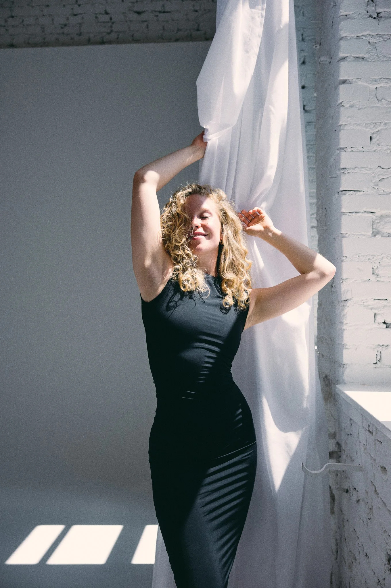 Eine Frau mit lockigem Haar trägt ein schwarzes Kleid und steht vor einem weißen Vorhang in einem hellen Raum mit weißen Wänden. Sie scheint sich zu amüsieren und posiert freudig.