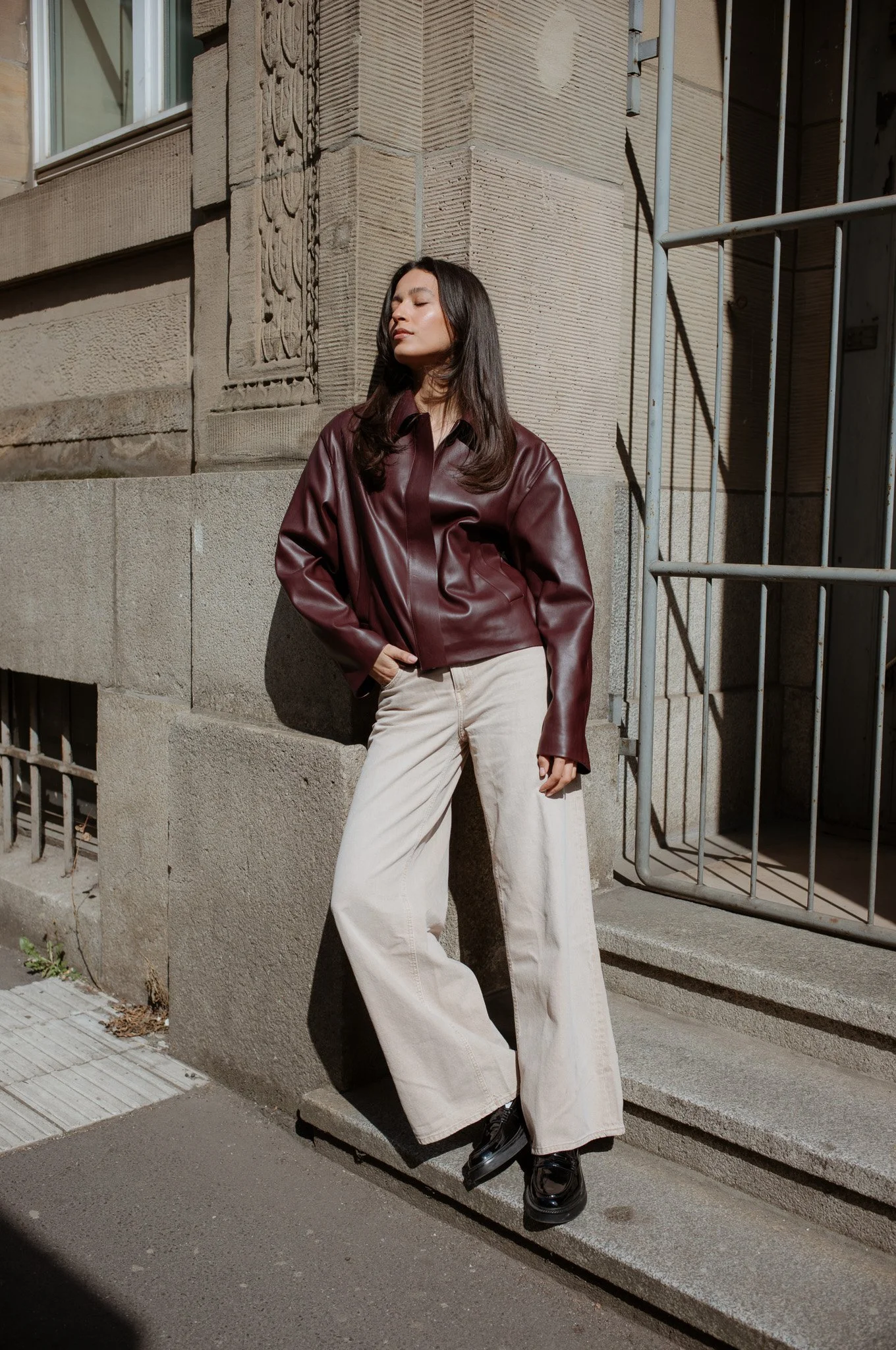Eine junge Frau lehnt an einer Steinmauer vor einem Gebäudefassade mit Fenster und Kunstrelief, trägt eine dunkelrote Lederjacke, helle weite Jeans und schwarze Schuhe, steht in der Sonne und schaut entspannt zur Seite.