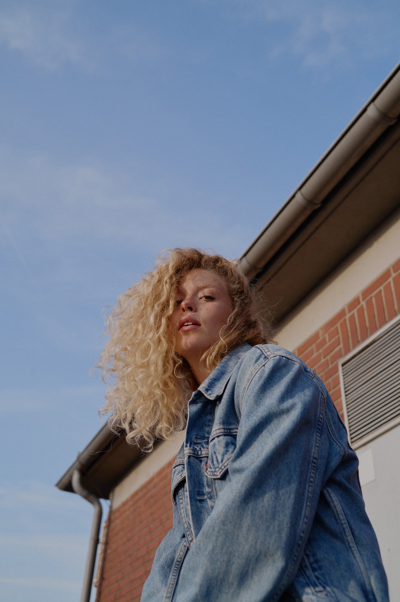 Junge Frau mit lockigem blonde Haar trägt eine Denim-Jacke vor einem Haus mit roten Ziegeln und einem blauen Himmel im Hintergrund.